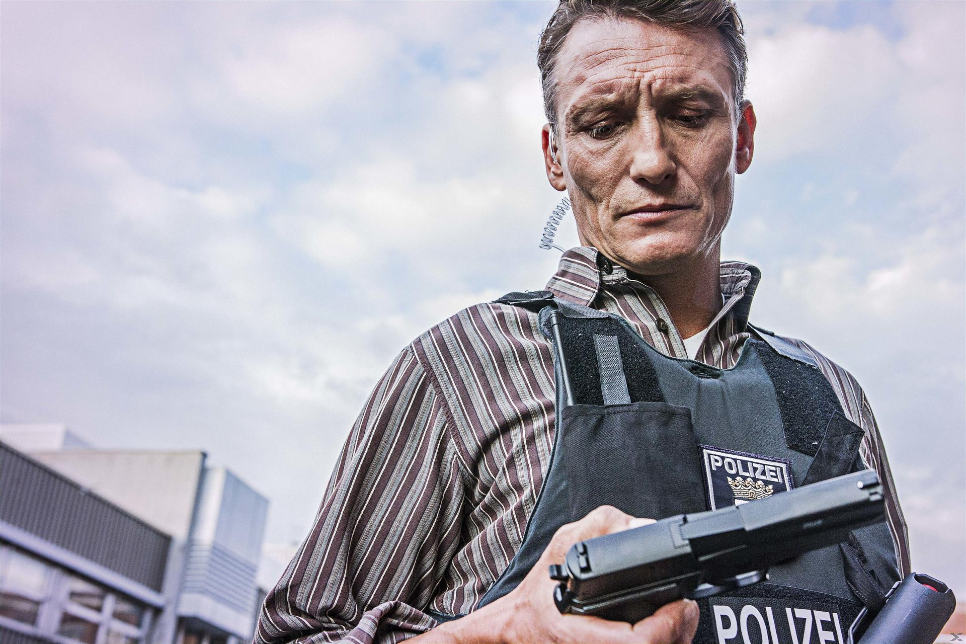 Ein Mann in gestreiftem Hemd und Weste hält eine Waffe. Das Wort POLIZEI ist sichtbar. Bewölkter Himmel im Hintergrund.