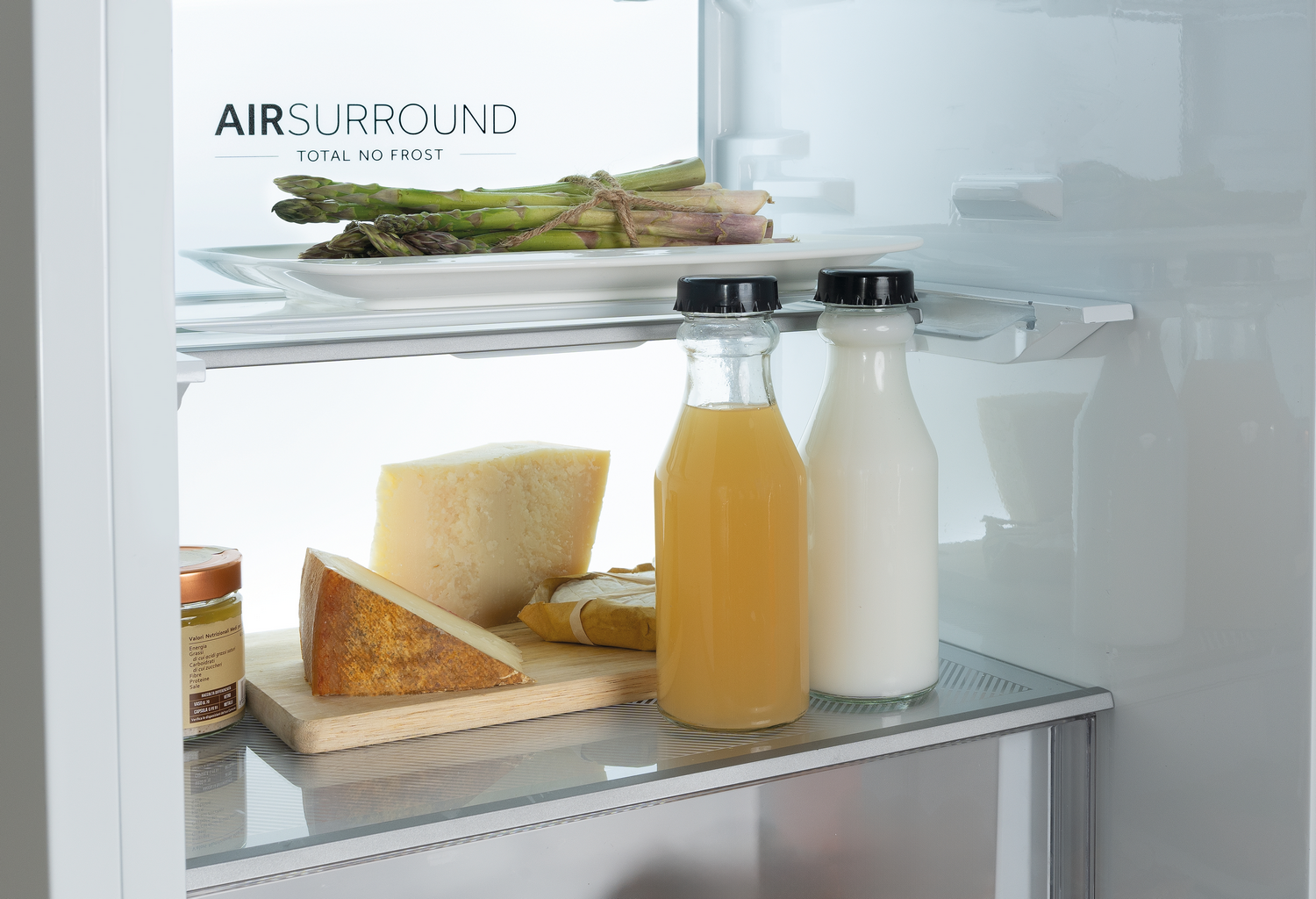 Dentro de un refrigerador: espárragos en un plato, queso, botellas de jugo y leche. El refrigerador está limpio y bien organizado.