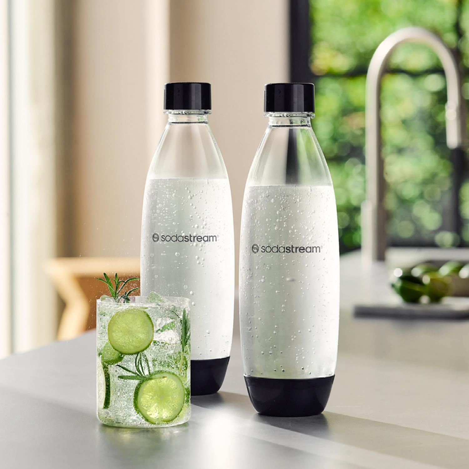 Due bottiglie SodaStream, un bicchiere con fette di lime ed erbe aromatiche e uno sfondo sfocato con una finestra e un lavandino.