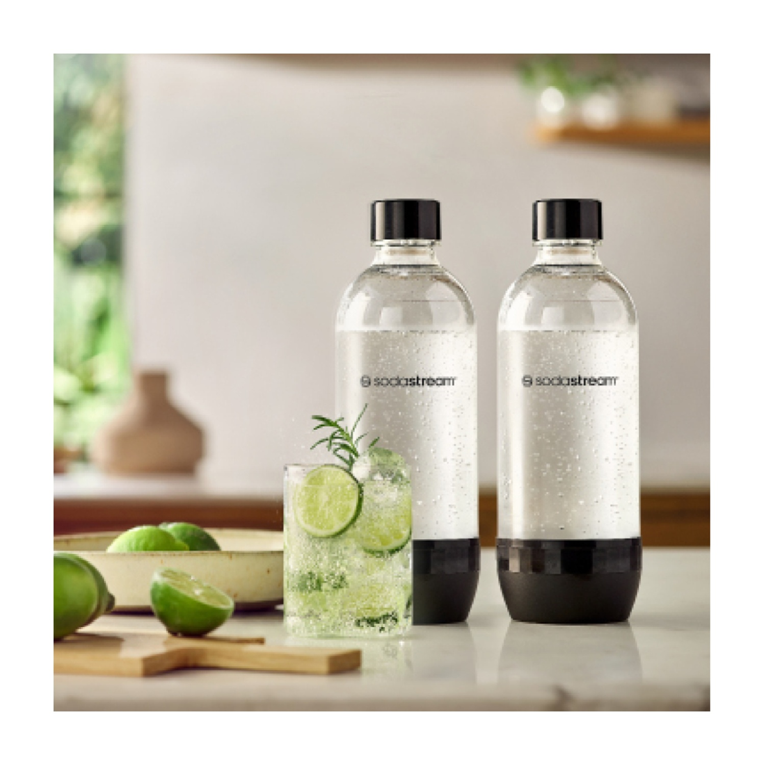 Due bottiglie SodaStream, un bicchiere di bevanda al lime e lime su un tagliere. Lo sfondo ha una finestra e una cucina sfocata.