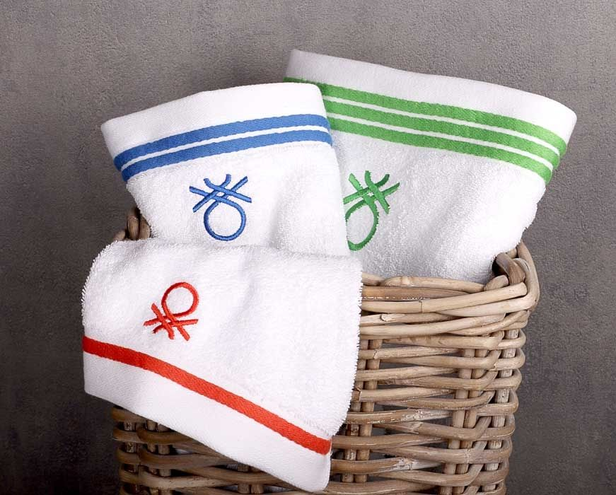 Három fehér törölköző színes csíkokkal és logókkal egy fonott kosárban. A csíkok kékek, zöldek és pirosak.