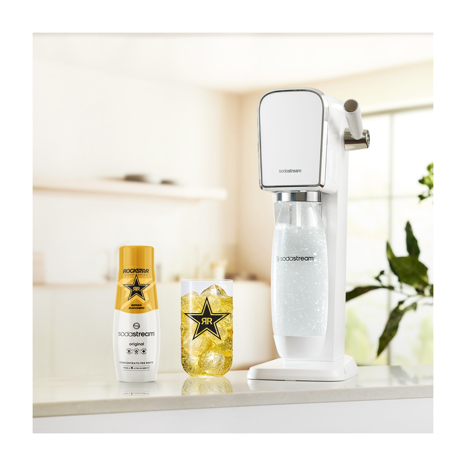 Una macchina SodaStream, un bicchiere di bevanda con ghiaccio e una bottiglia di energy drink Rockstar sono su un bancone bianco.