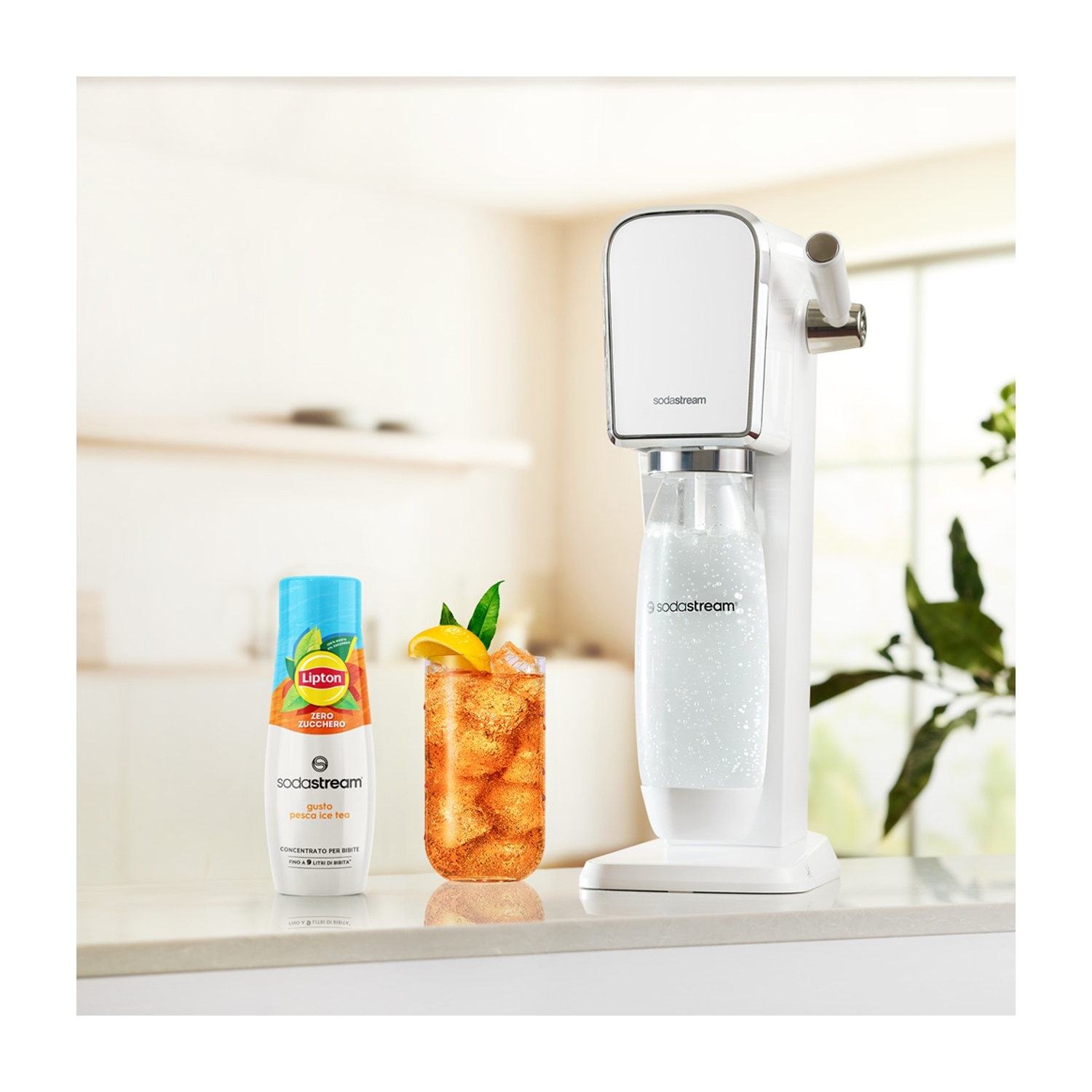 Una macchina SodaStream bianca, un bicchiere di tè freddo al limone e una bottiglia di sciroppo di tè freddo alla pesca Lipton sono su un bancone bianco.