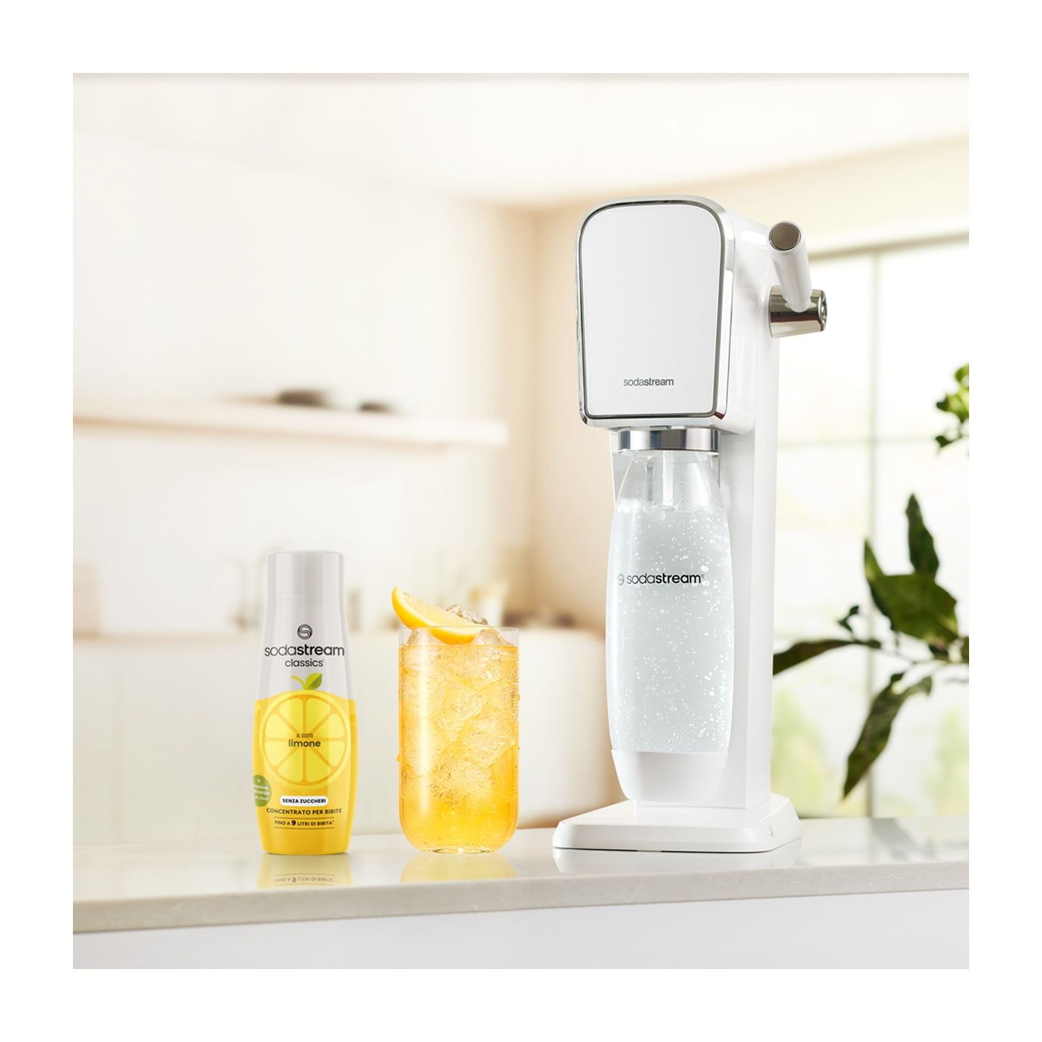 Una macchina SodaStream bianca con un bicchiere di limonata e una bottiglia di concentrato di limone su un piano di lavoro bianco.