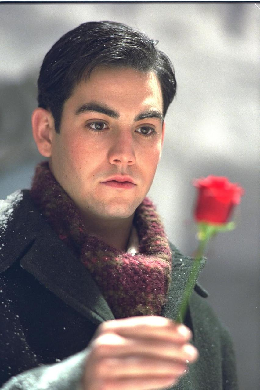 Ein Mann hält eine rote Rose. Er trägt einen Schal und einen Mantel. Schneeflocken sind auf seinem Haar und Mantel. Der Hintergrund ist verschwommen.