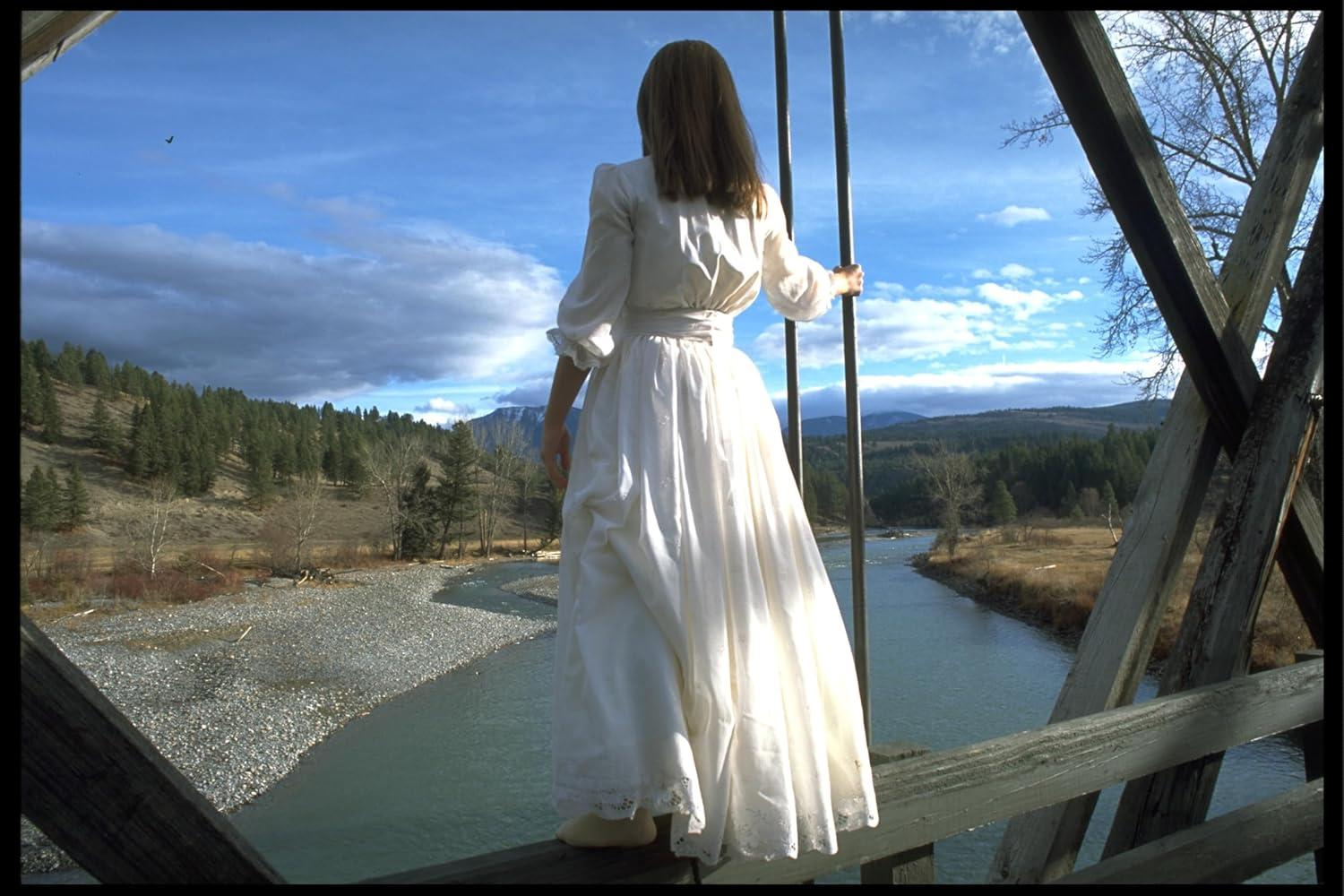 Eine Frau in einem weißen Kleid steht auf einer Holzbrücke und blickt auf einen Fluss und Berge. Der Himmel ist bewölkt.
