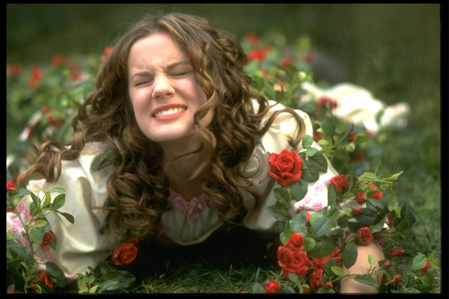 Eine Frau liegt im Gras, umgeben von roten Rosen, die Augen geschlossen, der Mund verzogen. Sie trägt ein cremefarbenes Kleid.