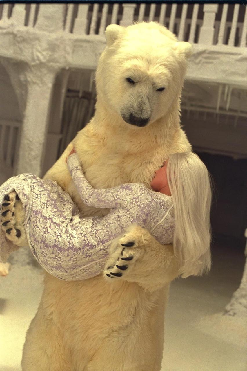 Ein Eisbär hält eine Frau im Arm. Sie trägt ein gemustertes Kleid und hat lange blonde Haare. Der Bär befindet sich in einer verschneiten Umgebung.