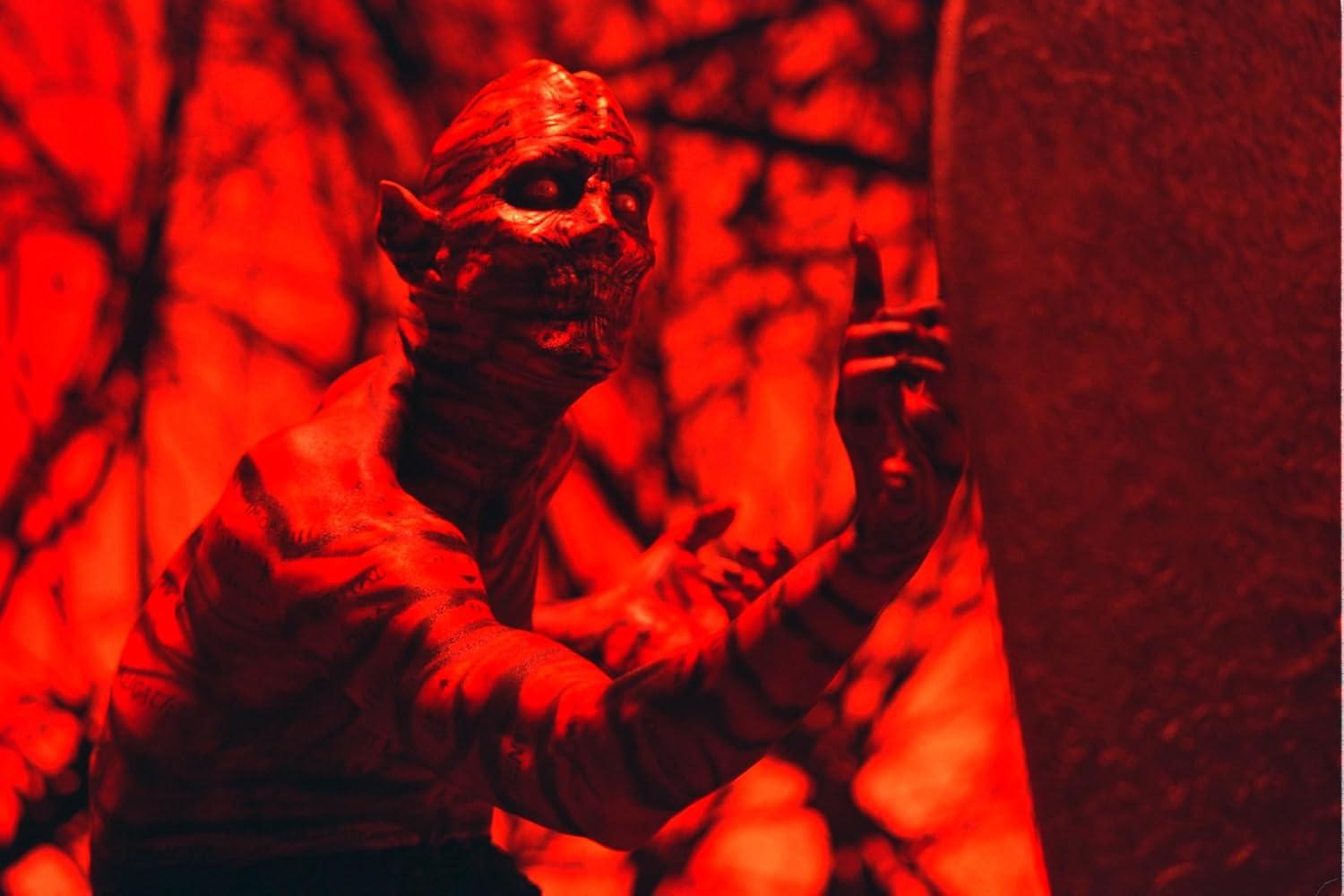 Ein rot getöntes Bild einer dämonischen Figur mit spitzen Ohren und gestreifter Haut, die nach oben zeigt. Der Hintergrund ist verschwommen rot.