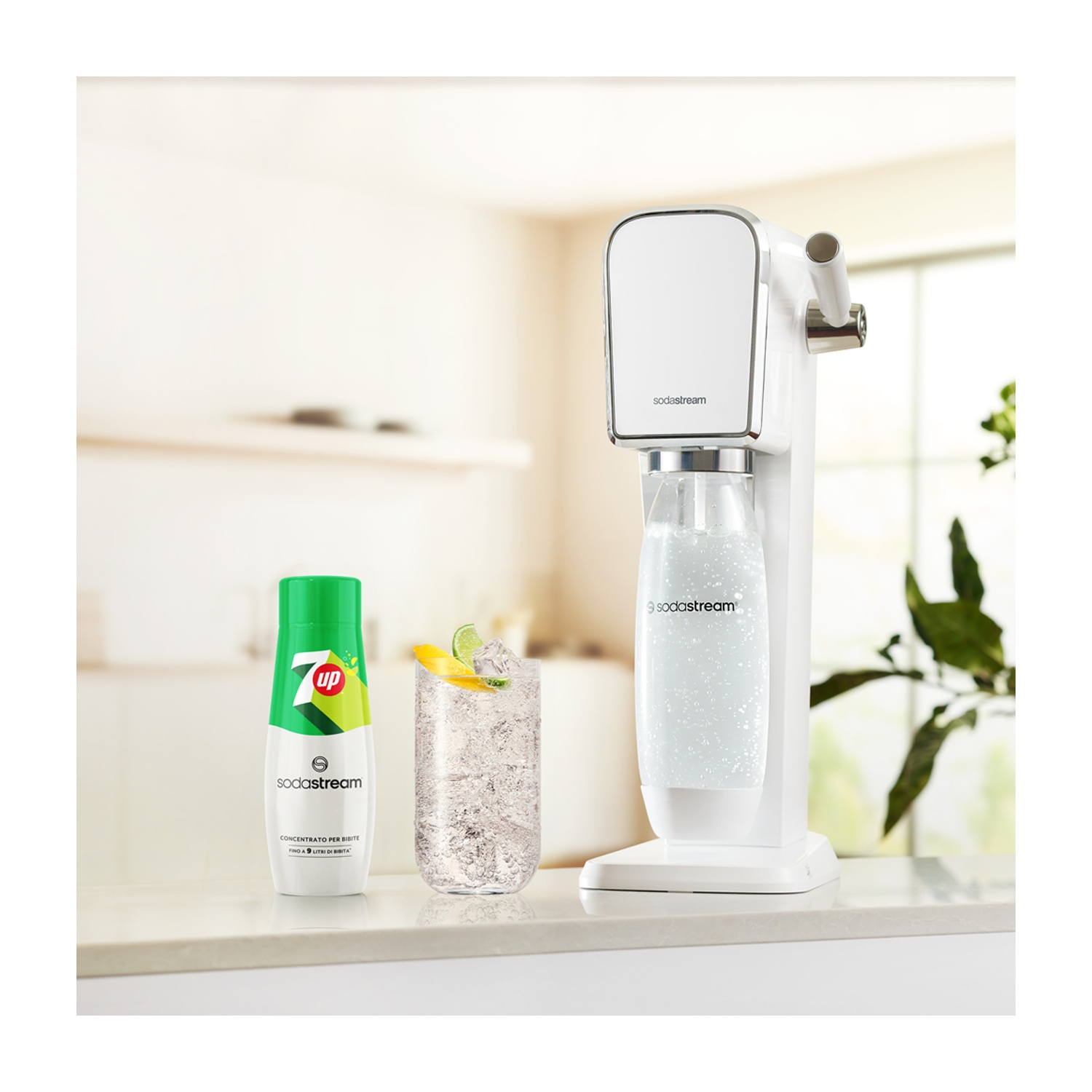 Una macchina SodaStream bianca, una bottiglia di 7up, un bicchiere con ghiaccio e limone e una pianta sullo sfondo. Tutto su un piano bianco.
