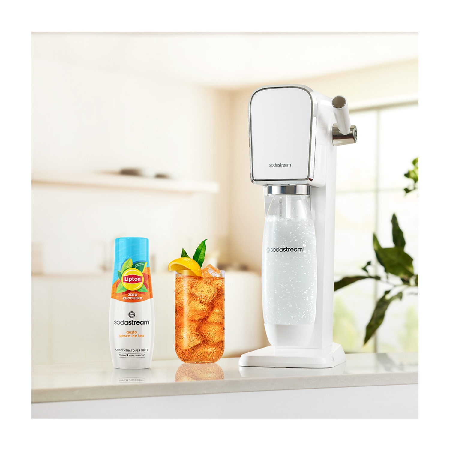Una macchina SodaStream bianca con un bicchiere di tè freddo, una bottiglia di Lipton e un bicchiere di soda. Tutto su una superficie bianca.