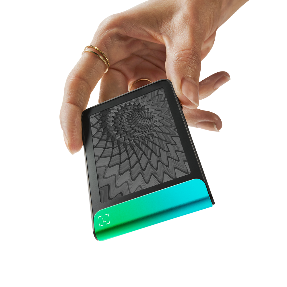 Una mano sostiene un dispositivo negro con una base degradada verde-azul. La pantalla muestra un patrón gris y arremolinado. Anillos adornan los dedos.