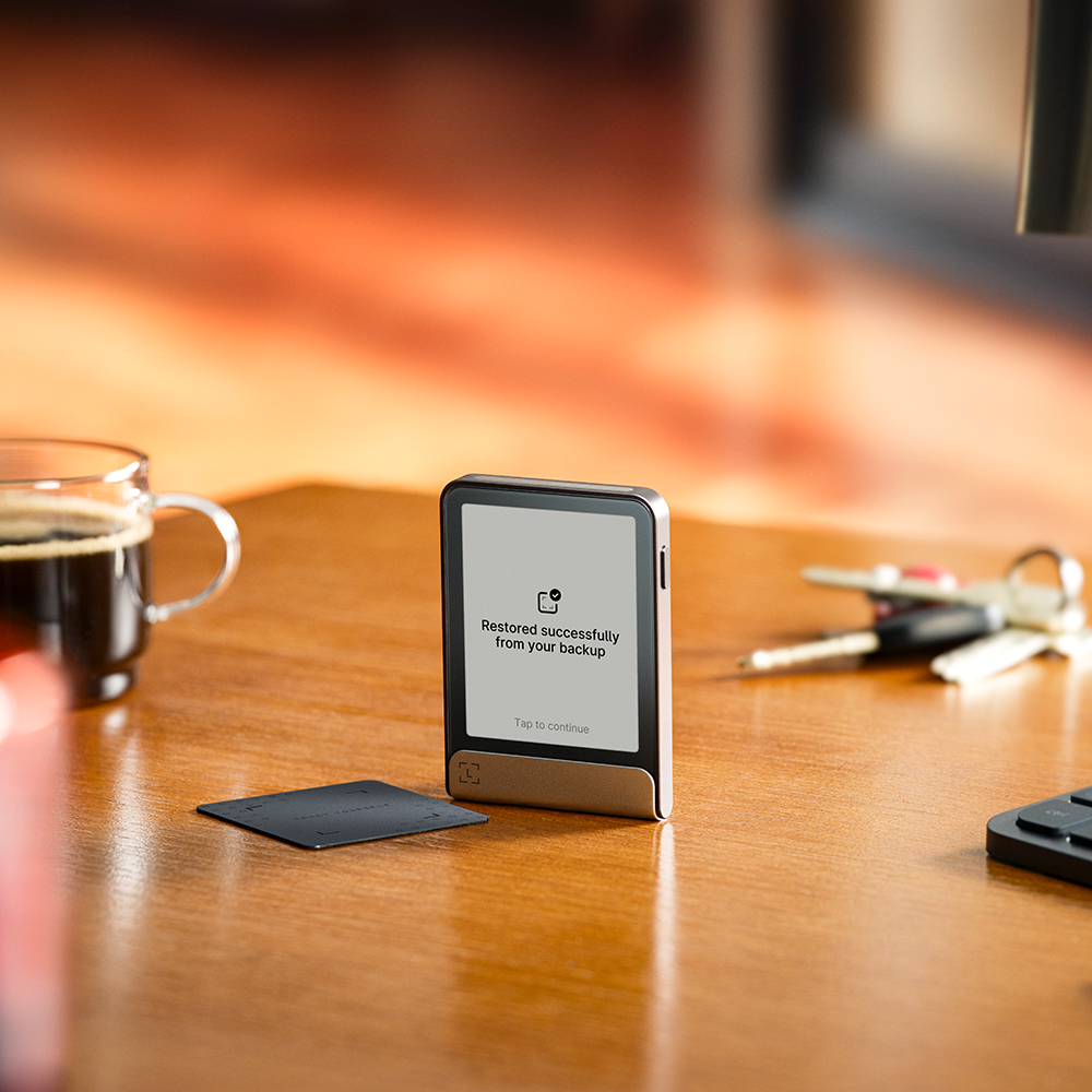 Un dispositivo inteligente con pantalla que muestra 'Restaurado correctamente'. Tarjeta de crédito, llaves y taza de café sobre mesa de madera.