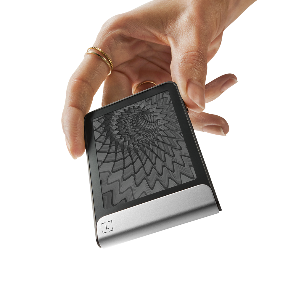 Una mano sostiene un pequeño dispositivo negro y plateado que muestra un patrón de espiral gris. La mano lleva un anillo de oro.