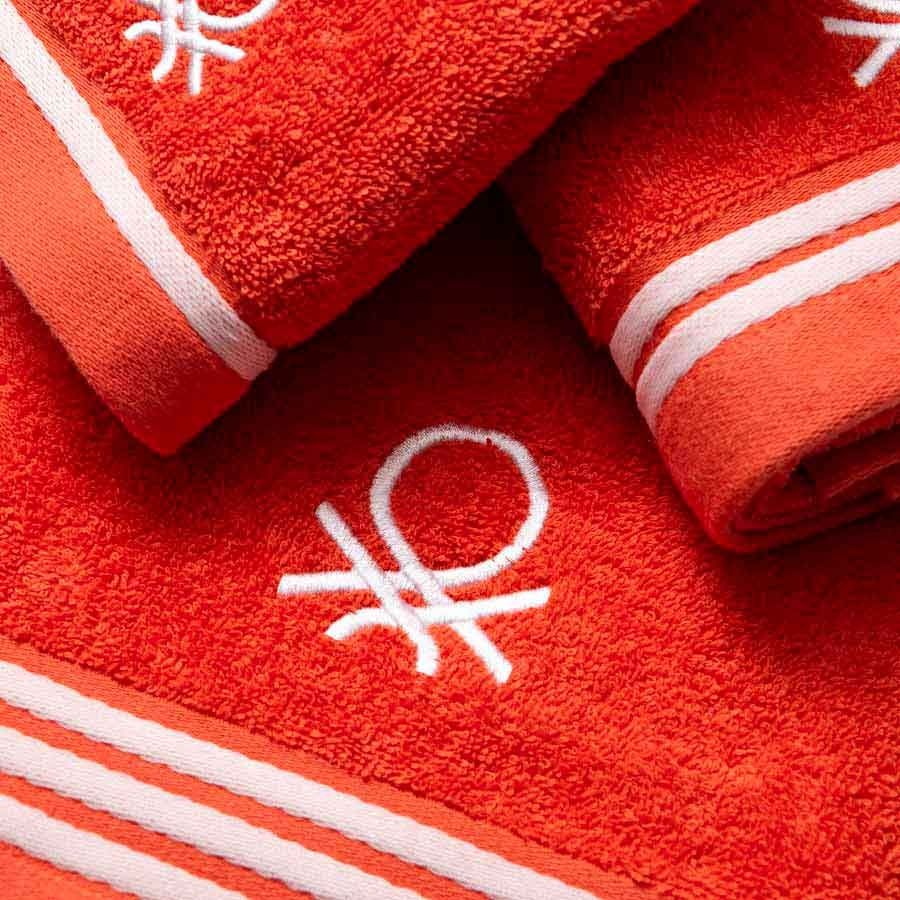 Narancssárga törölközők közelképe fehér csíkokkal és fehér logóval. A törölközők össze vannak hajtva és egymásra vannak rakva.
