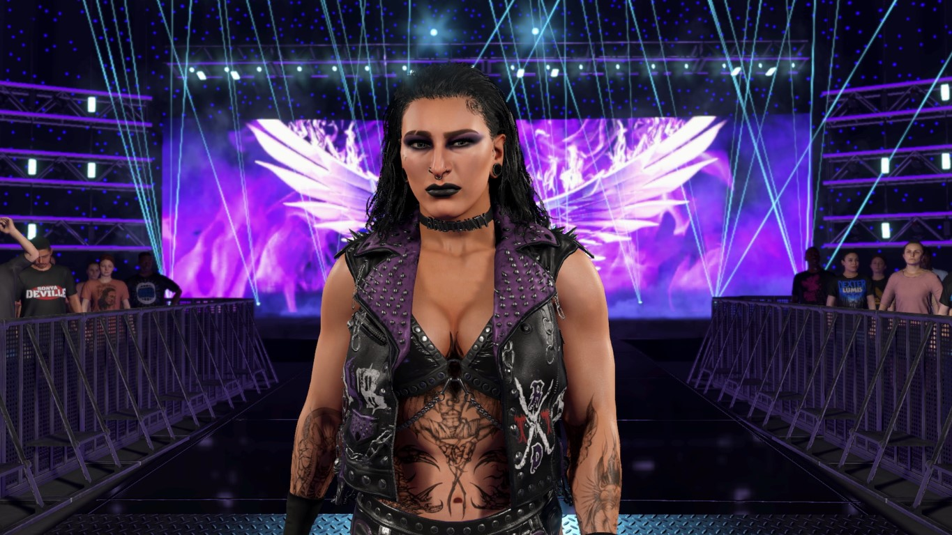 Kobieta z ciemnym makijażem i tatuażami stoi na scenie. Nosi czarno-fioletowy strój, choker i czarną szminkę.