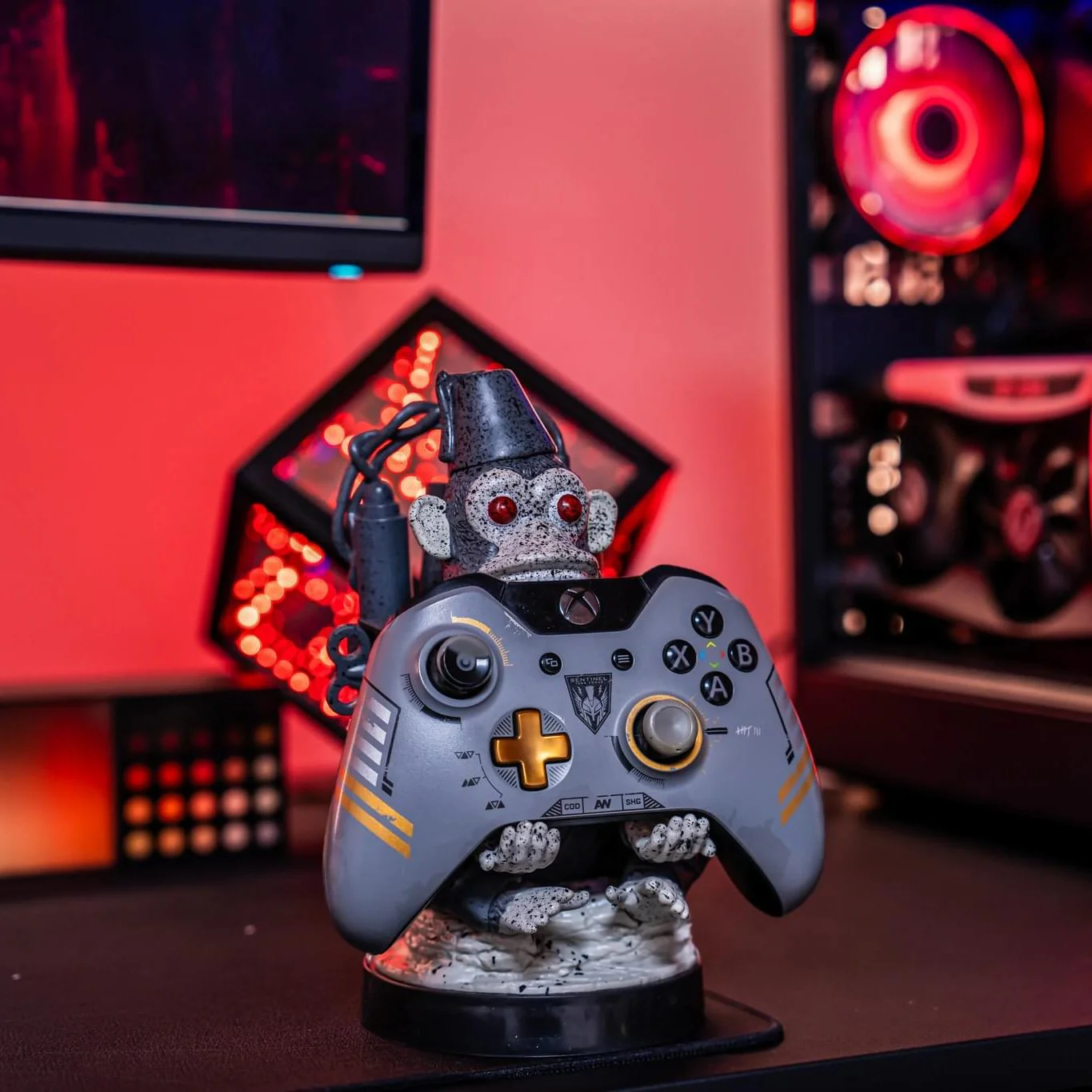 Un supporto per controller Xbox a tema scimmia. Il controller è grigio con dettagli arancioni. Lo sfondo include un computer e illuminazione rossa.