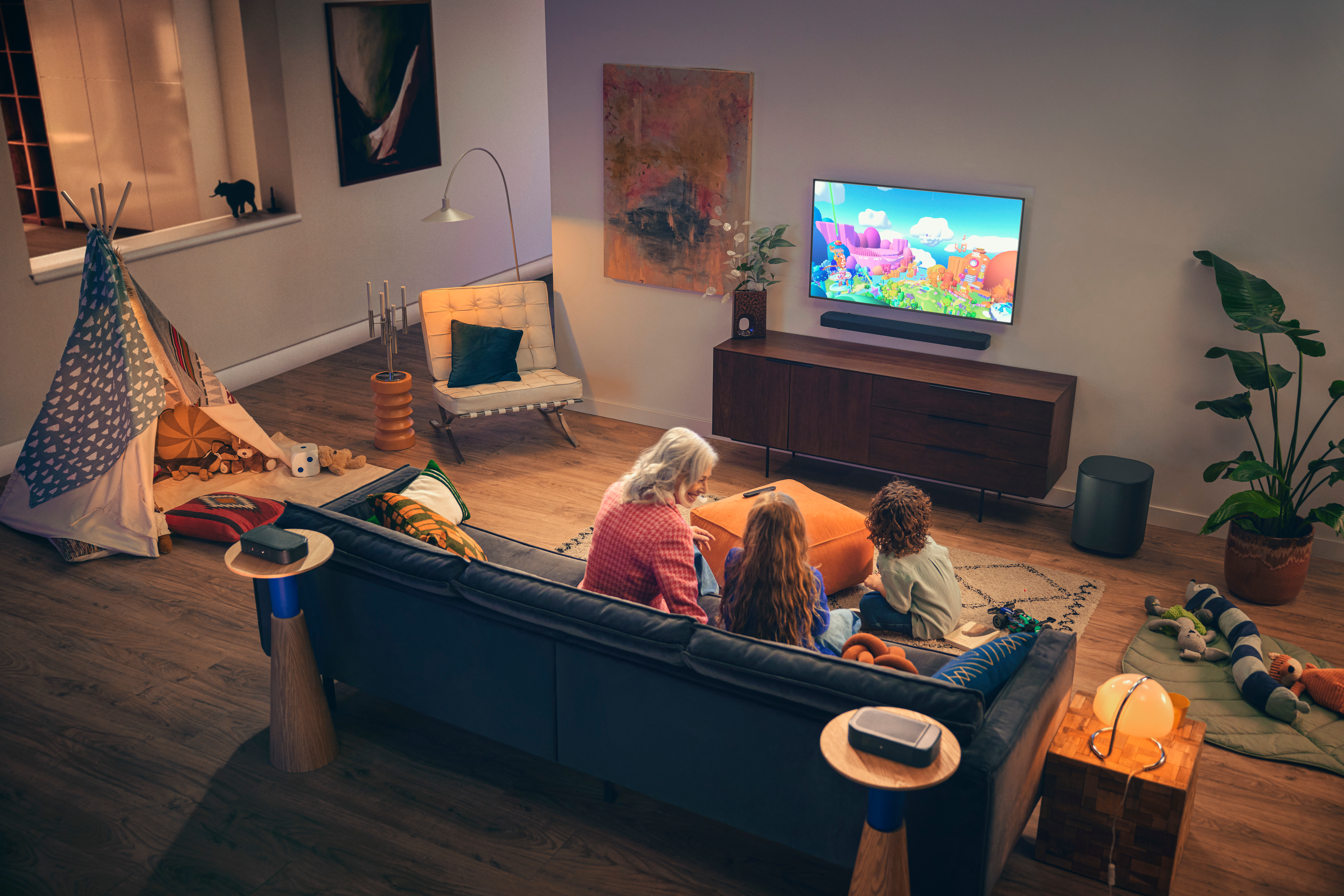 Eine Familie schaut fern. Ein Zelt steht am Fenster. Ein Sofa und ein Holzschrank sind im Bild.