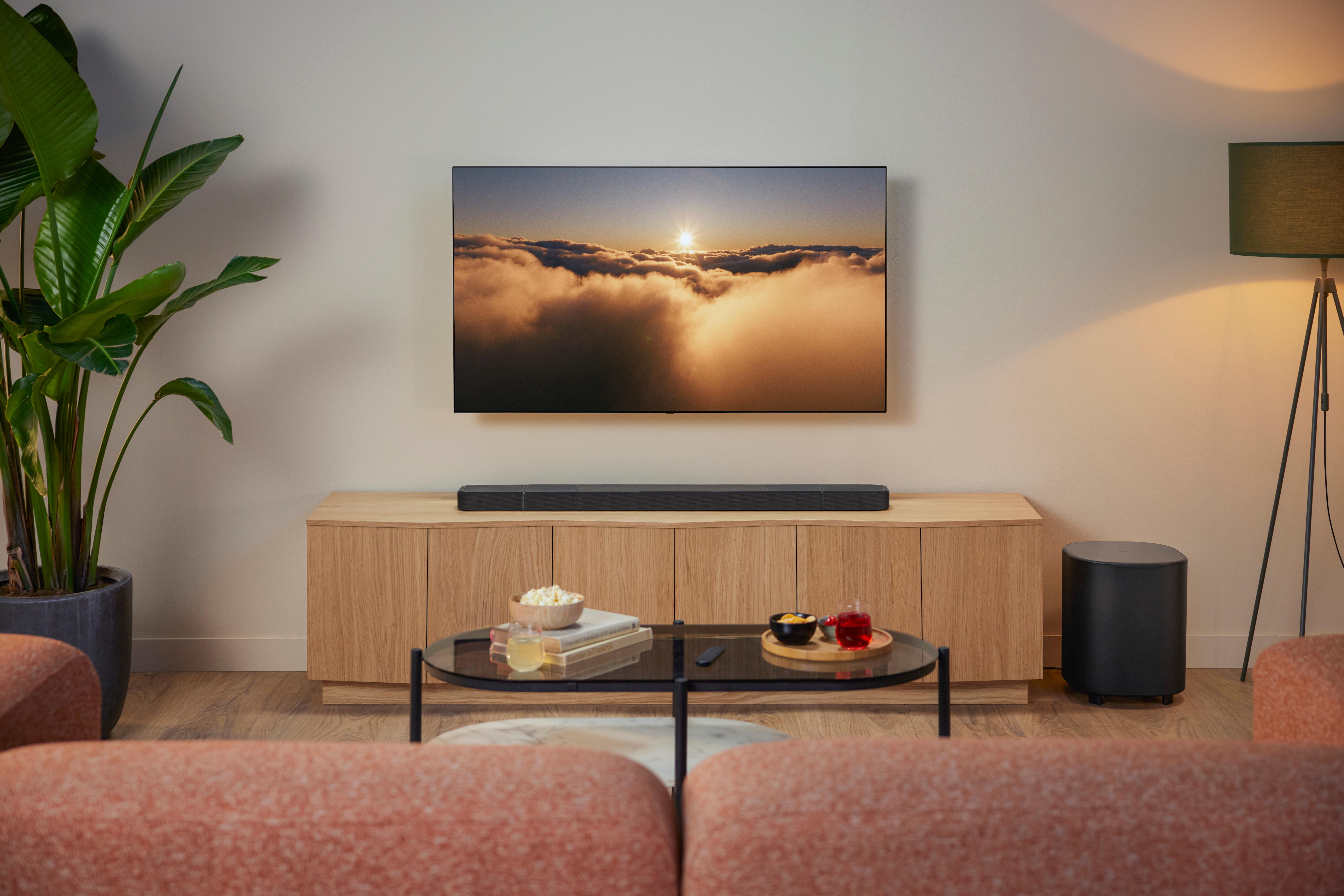 Wohnzimmerszene mit Fernseher, Soundbar, Holzschrank, Couchtisch mit Snacks, Sofa, Stehlampe und einer großen Pflanze.