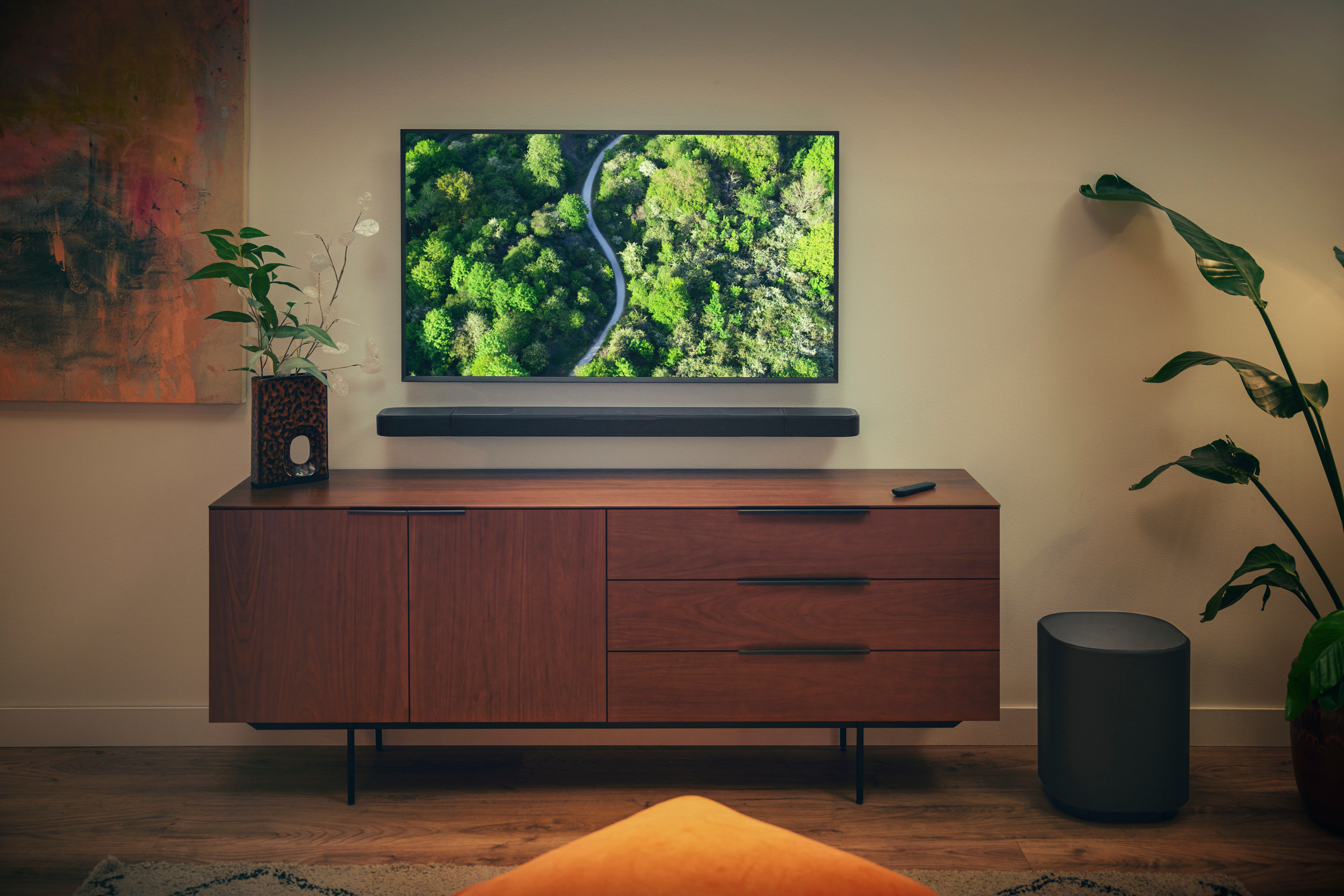 Ein Wohnzimmer mit einem Fernseher, der einen Waldweg zeigt, über einem Holzschrank. Eine Soundbar, Pflanze und Lautsprecher sind vorhanden.