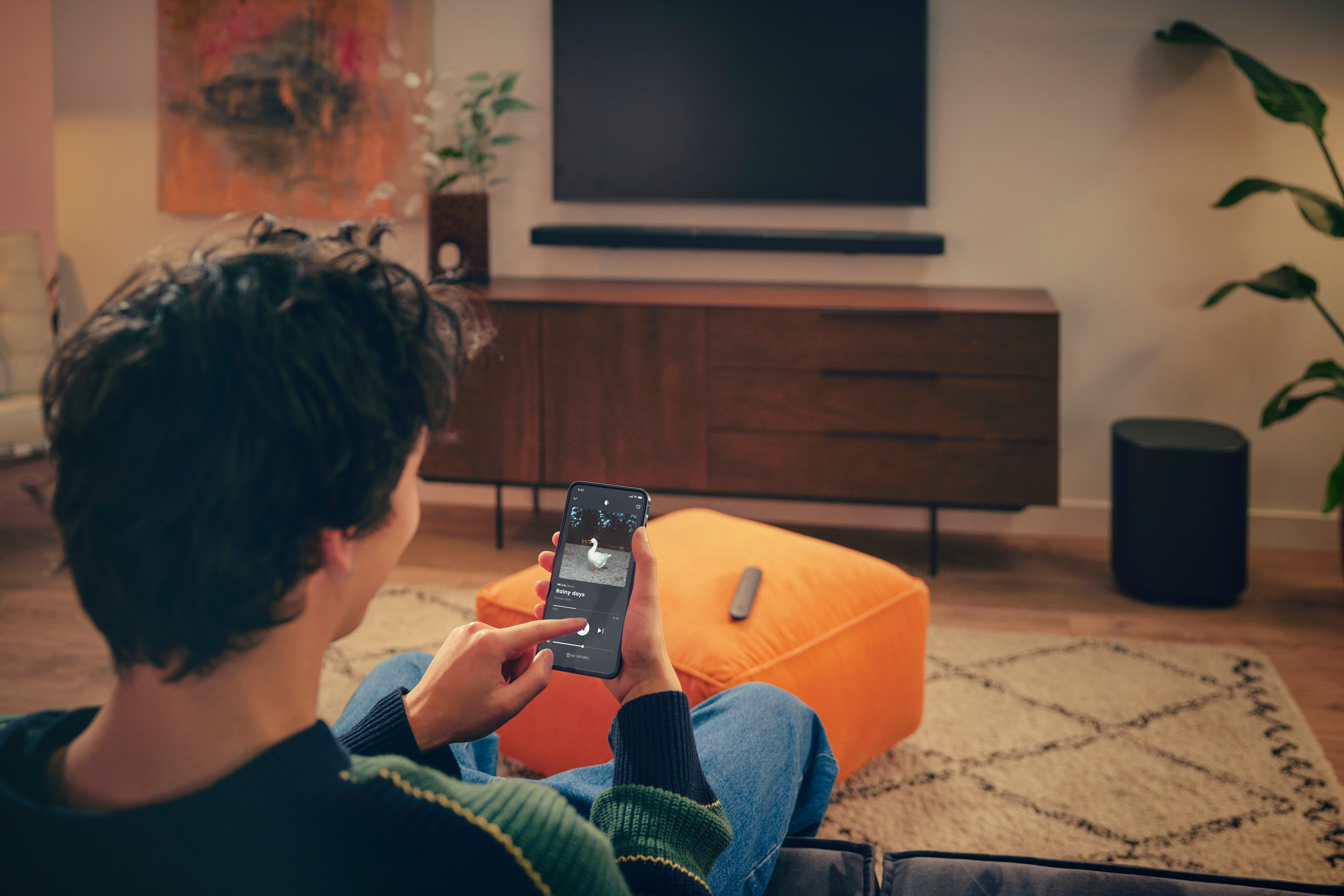 Eine Person benutzt ein Smartphone, um Musik zu steuern. Ein orangefarbener Hocker, ein Fernseher und Lautsprecher befinden sich im Hintergrund.