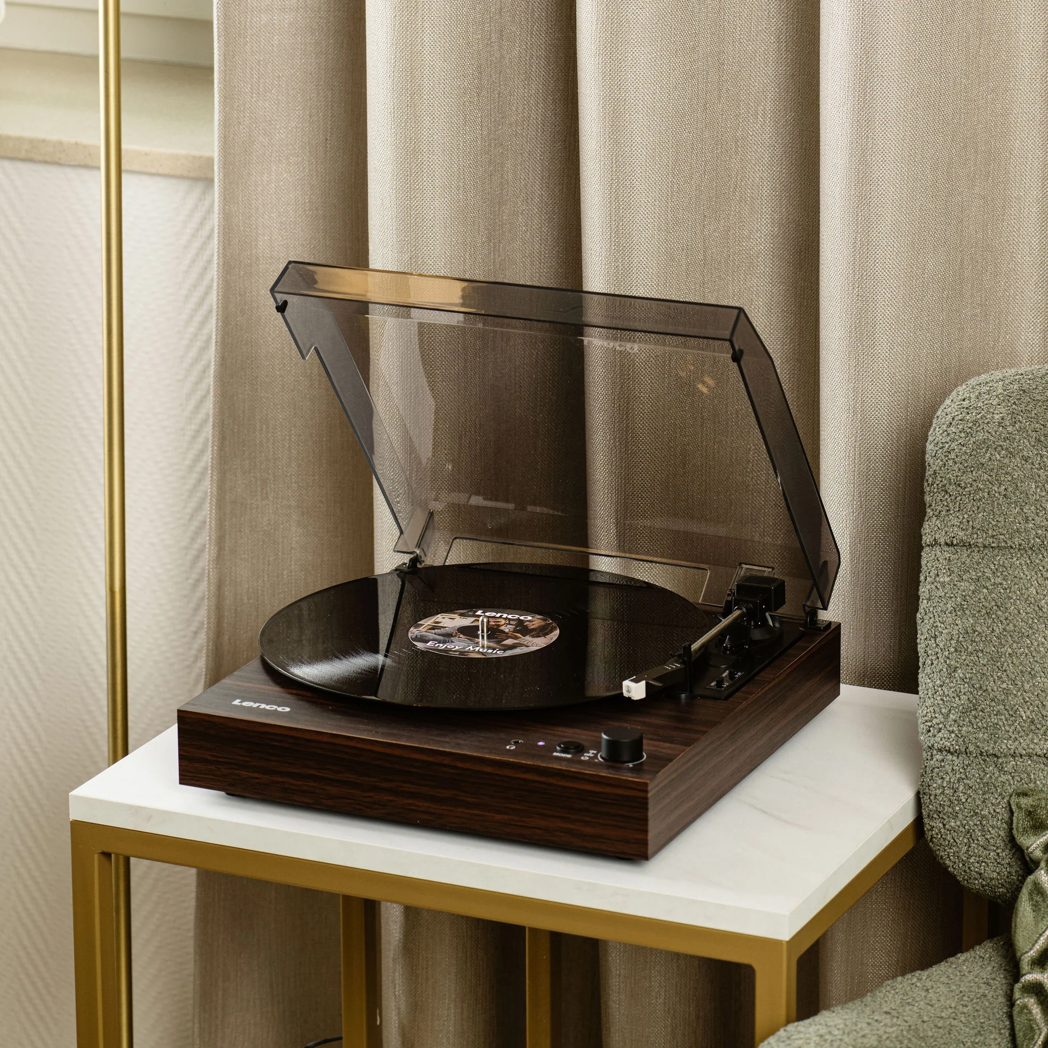 Une platine avec un disque est sur une table blanche et dorée. Derrière, une chaise verte et des rideaux beiges.