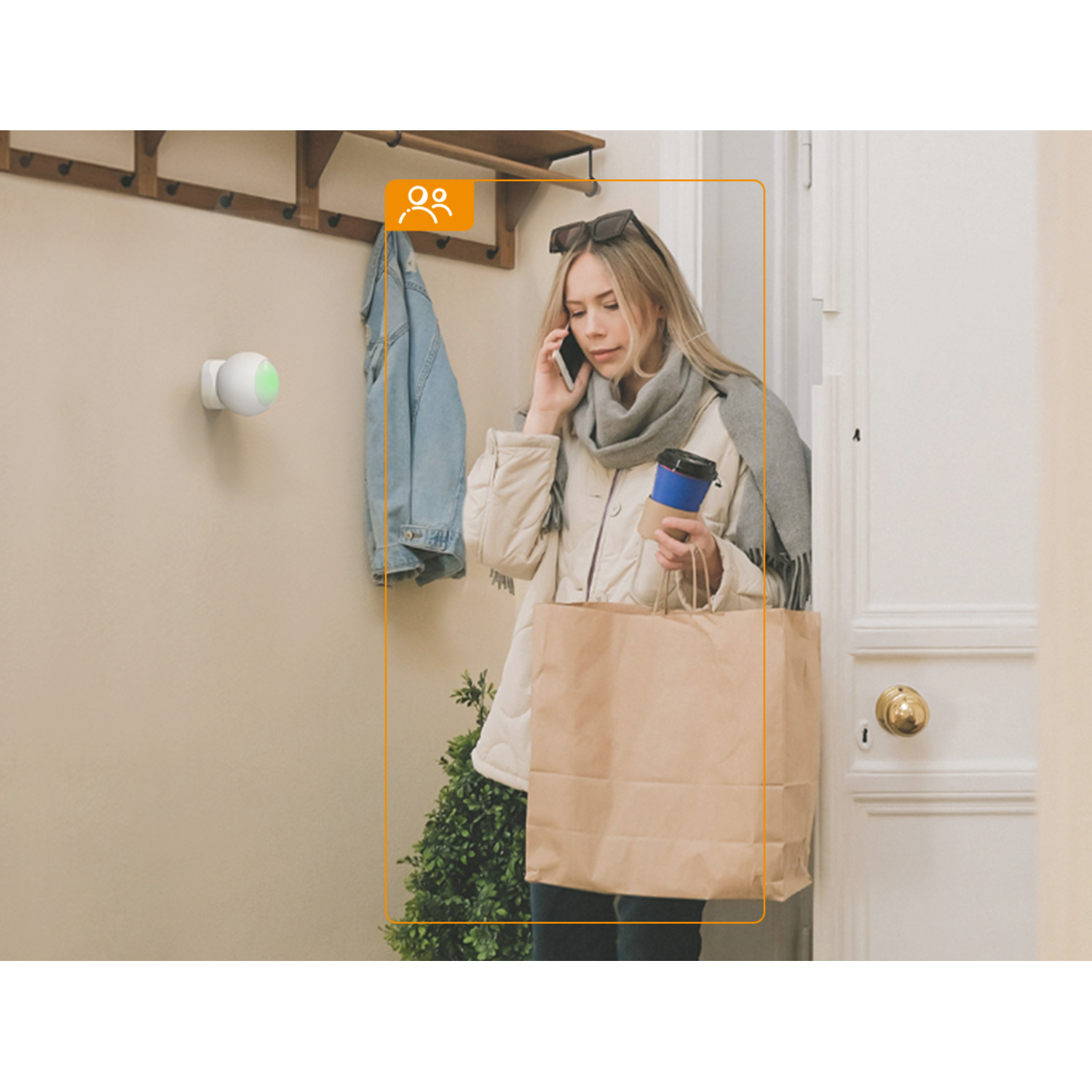 A woman talking on the phone, carrying a bag and coffee. A security camera and a coat rack are on the wall. Open door.