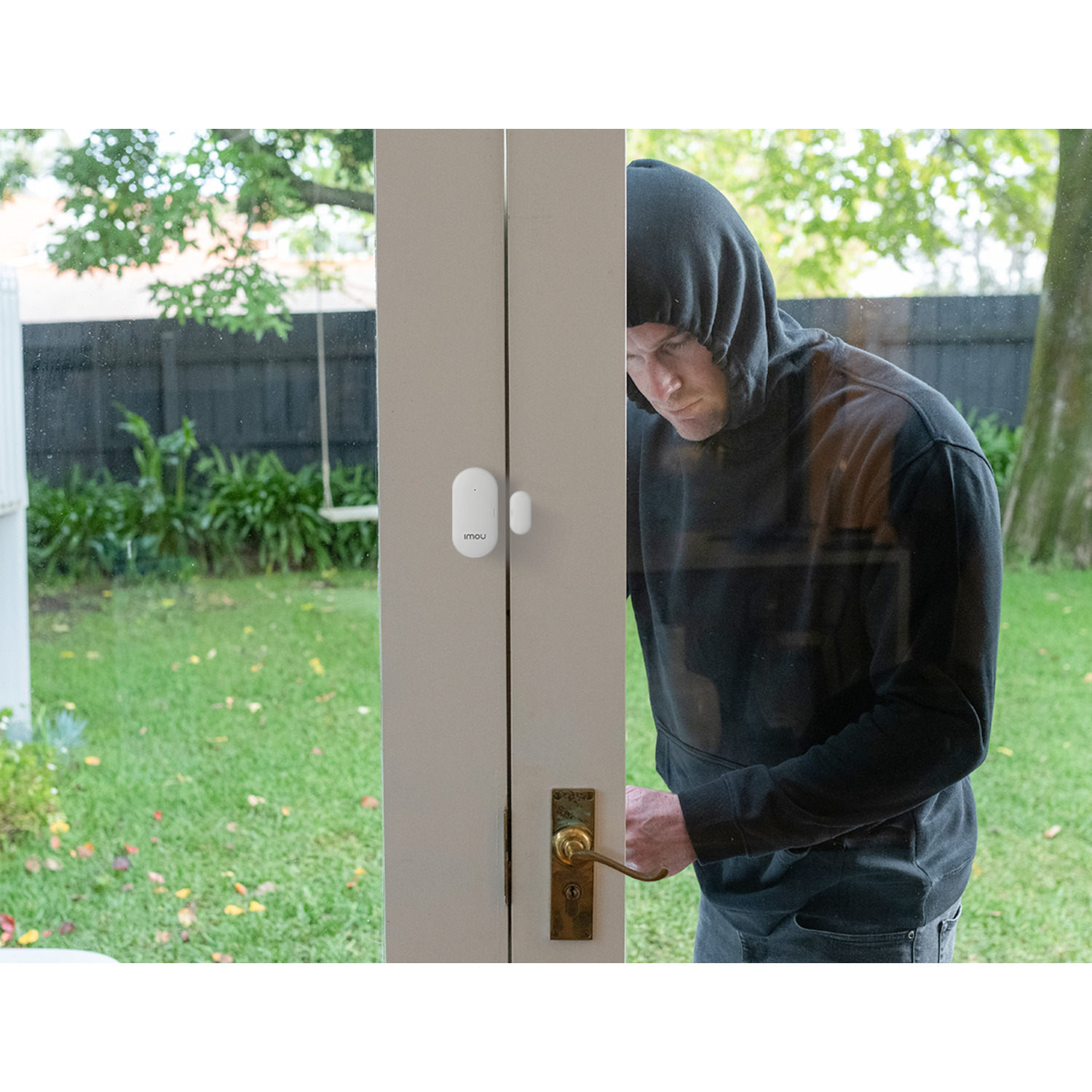 A man in a black hoodie attempts to open a door. A white security sensor is attached. The background shows a yard with a tree.
