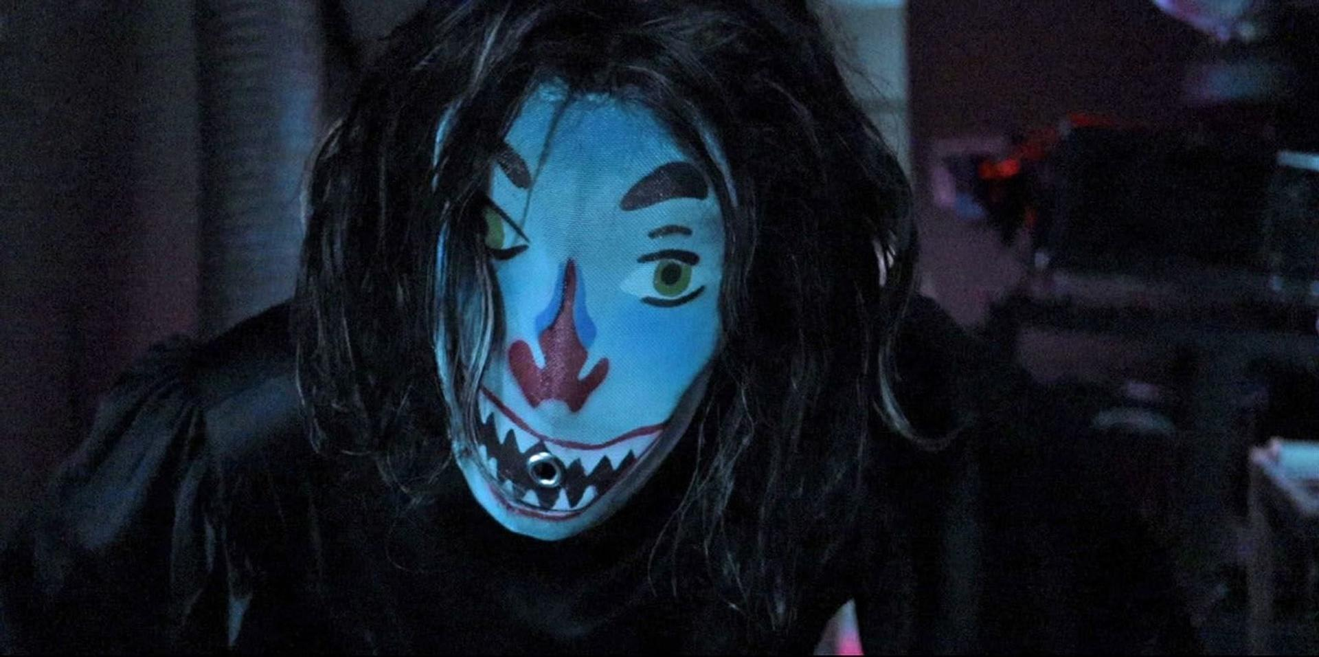 Eine Person mit einer blauen Gesichtsmaske mit clownartigem Design und schwarzem Haar. Ein dunkler Hintergrund mit einigen Lichtern.