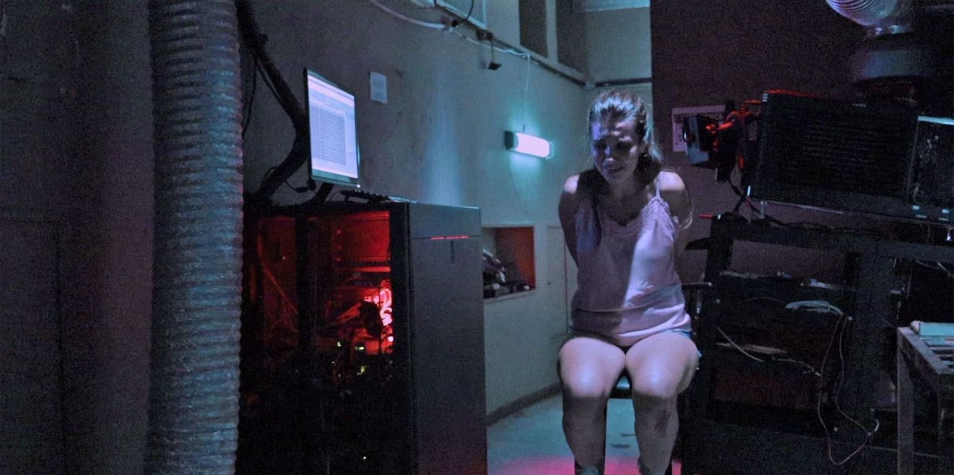 Eine Frau ist in einem Keller an einen Stuhl gefesselt. Ein Computer und ein Projektor sind sichtbar. Der Raum ist spärlich beleuchtet.