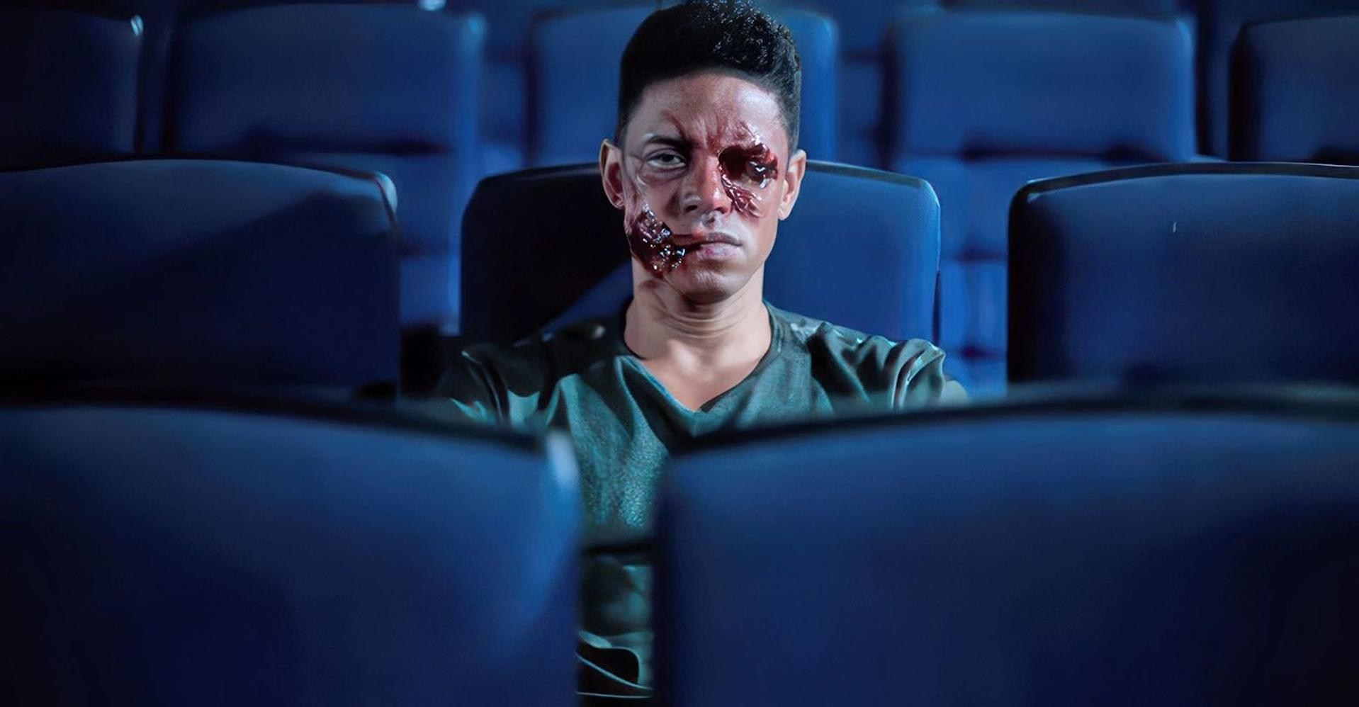 Ein Mann mit Gesichtsverletzung sitzt in einem Kino. Der Hintergrund zeigt blaue Kinosessel.