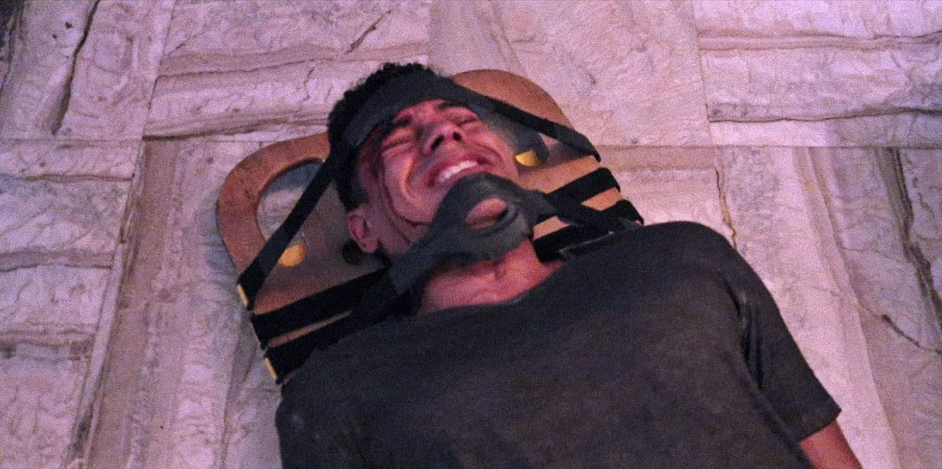 Ein Mann liegt gefesselt an einem Holzrahmen und trägt eine Maske. Sein Gesicht zeigt Not, und es ist Blut zu sehen. Der Hintergrund ist Stein.