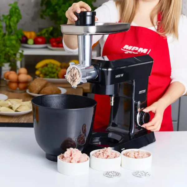 Kobieta mieli mięso czarną maszyną kuchenną MPM. Na białym stole miski z mielonym mięsem i składnikami.