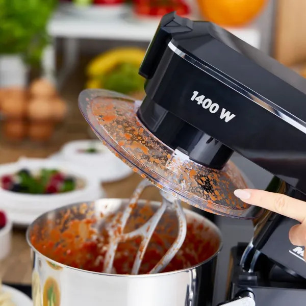 Czarny robot kuchenny z przezroczystą pokrywą, wewnątrz czerwony sos. Ręka otwiera pokrywę. W tle: owoce i warzywa.