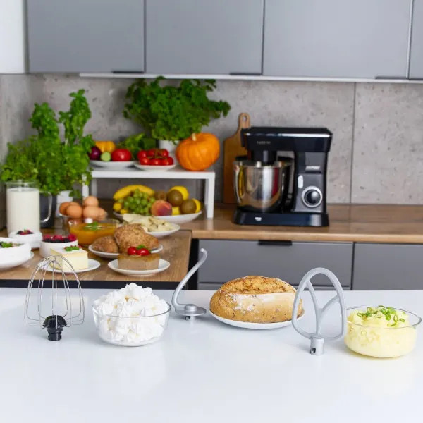 Scena kuchenna z czarnym mikserem, składnikami, owocami, chlebem i przyborami na białym stole. W tle szafki.