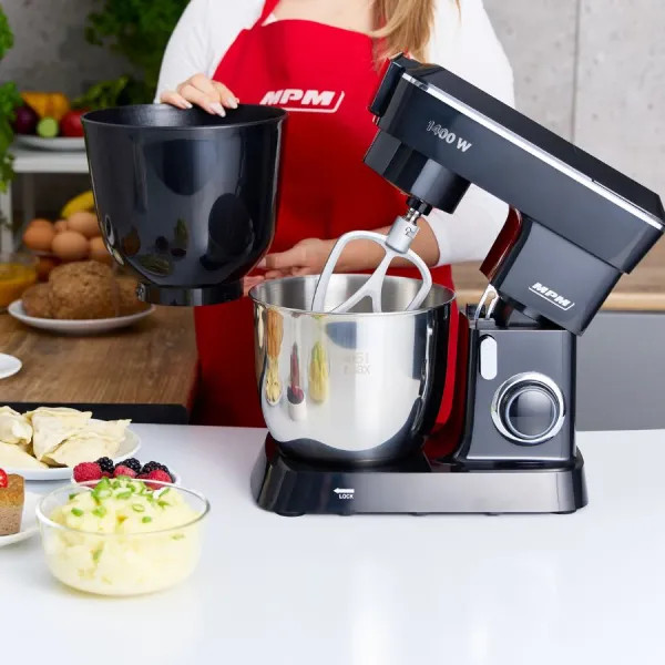 Kobieta w czerwonym fartuchu trzyma czarną miskę nad mikserem. Na stole puree ziemniaczane i jagody.