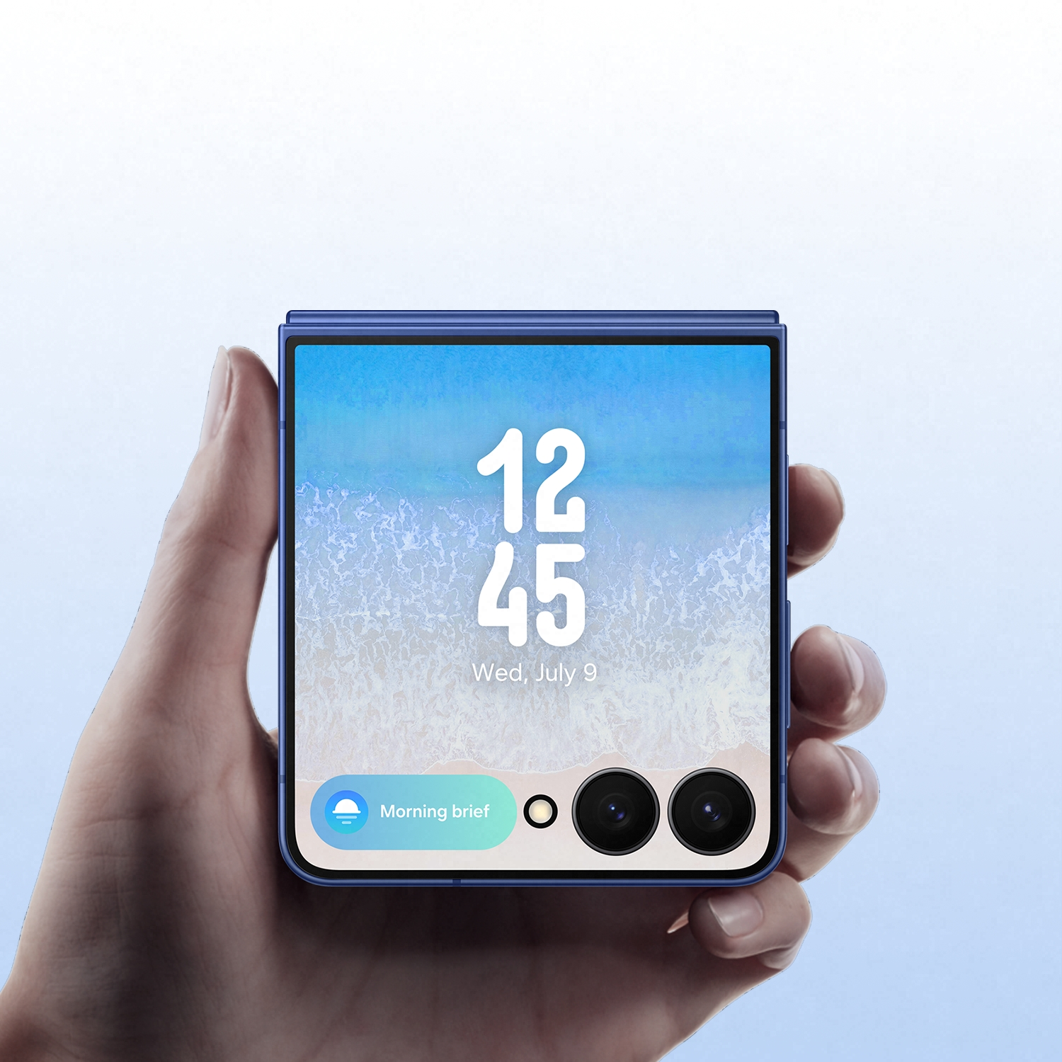Una mano sostiene un teléfono azul plegable. La pantalla muestra la hora, 12:45, y un fondo de pantalla de playa. Abajo hay iconos de cámara y notificación.