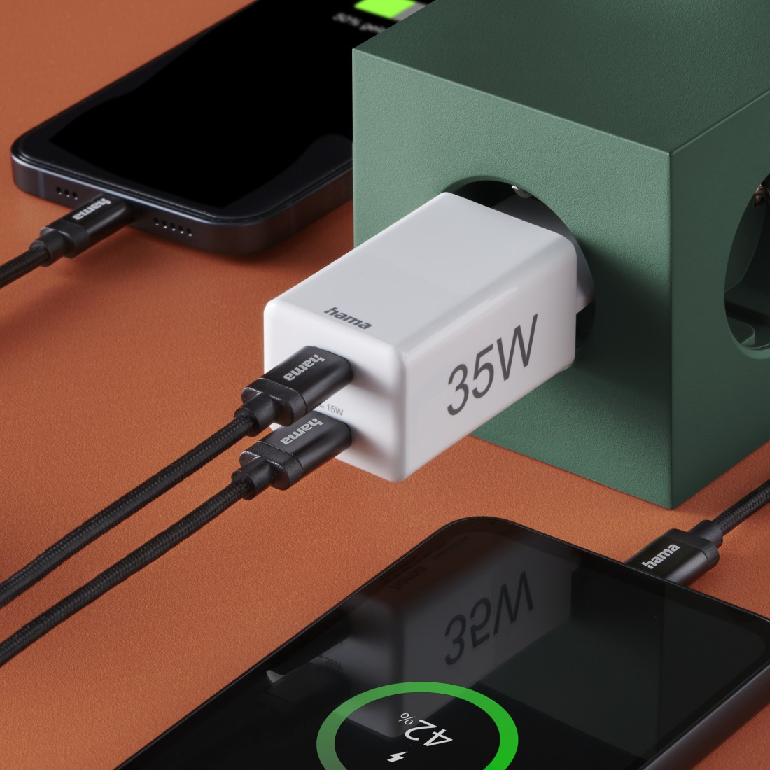 Weißes Hama-Ladegerät mit zwei schwarzen Kabeln, die an Handys angeschlossen sind. Ein Telefon lädt, zeigt 42 %. Das Ladegerät ist 35 W.