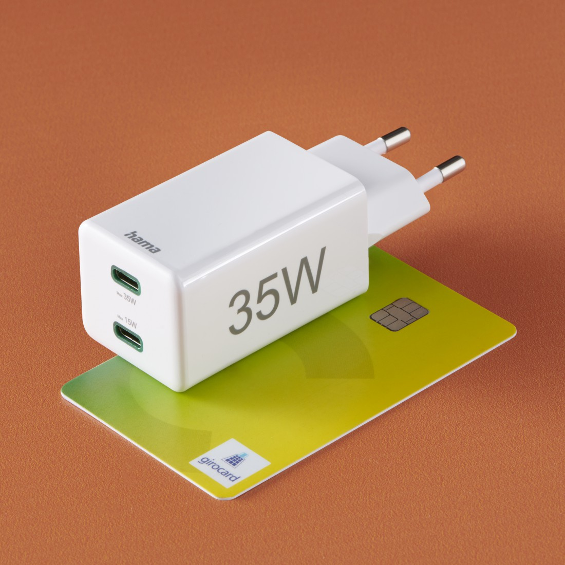 Ein weißes 35-W-Ladegerät mit einer Kreditkarte auf orangefarbenem Untergrund. Das Ladegerät hat zwei USB-C-Anschlüsse und das Wort „hama“.