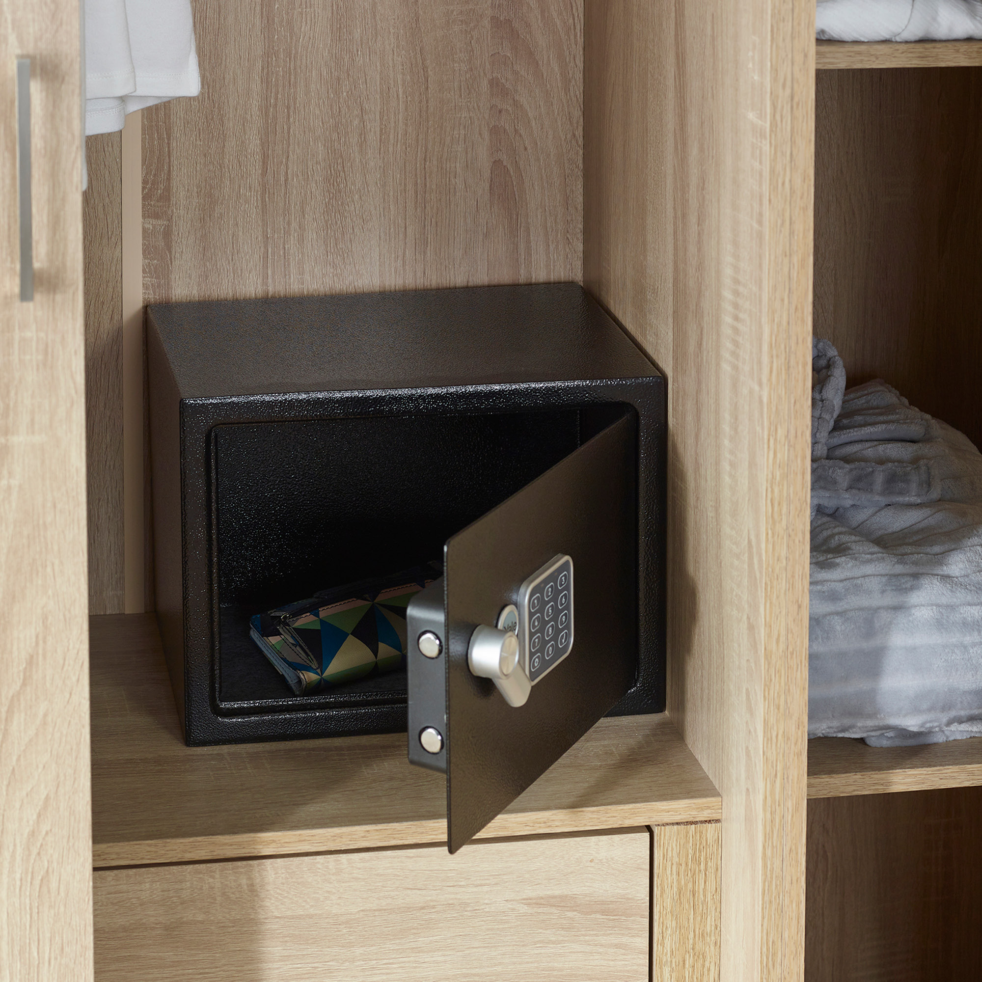 Ein schwarzer Safe mit offener Tür in einem Holzschrank. Im Inneren befindet sich eine Brieftasche. Handtücher und Kleidung sind ebenfalls sichtbar.