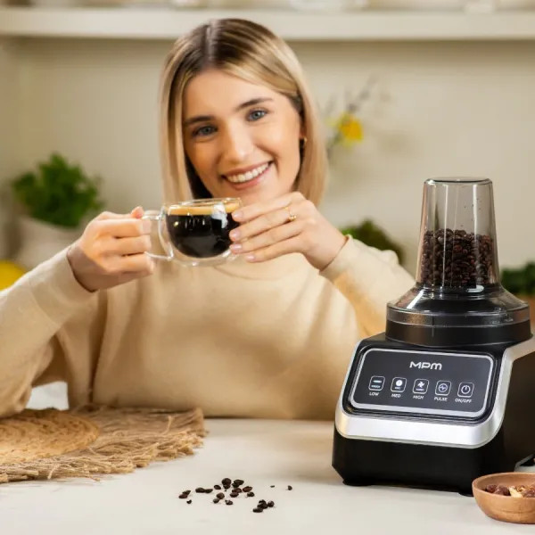 Kobieta trzymająca kawę, uśmiechnięta. Młynek do kawy i ziarna na stole. Tło kuchni.
