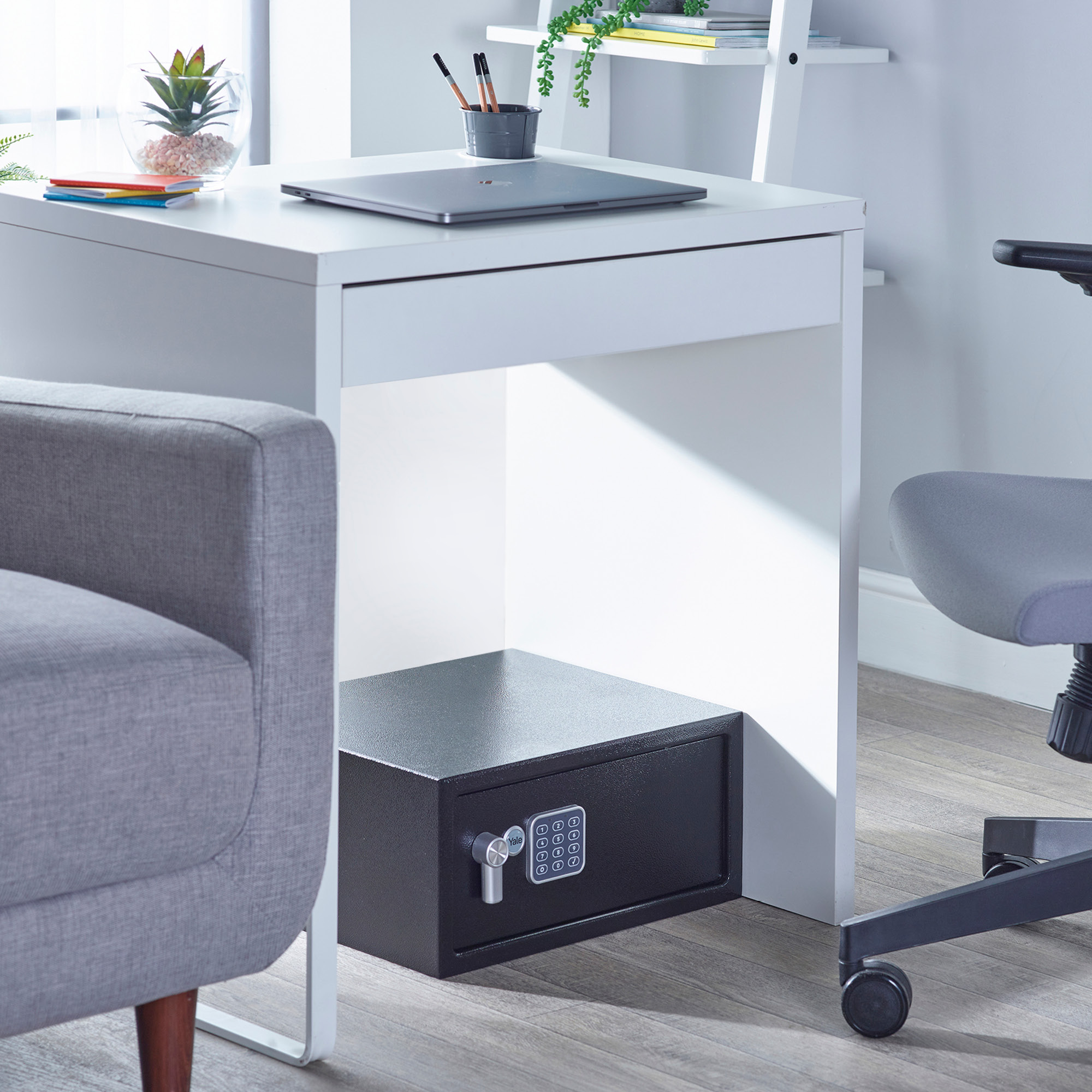 Ein schwarzer Safe unter einem weißen Schreibtisch. Auf dem Schreibtisch steht ein Laptop. Rechts steht ein grauer Stuhl. Links ein grauer Sessel.