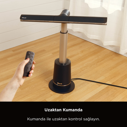 Siyah ve altın renkli bir vantilatör ahşap zeminde duruyor. Bir el uzaktan kumandayı tutuyor. Arka planda beyaz bir perde var.