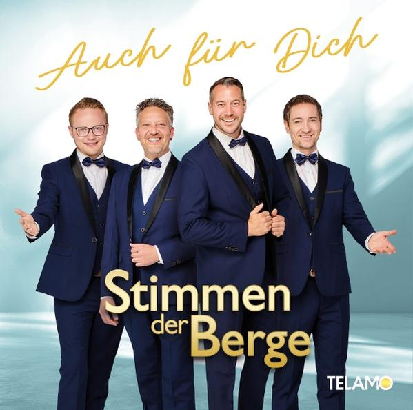 Vier Männer in Anzügen posieren. Oben steht 'Auch für Dich', unten 'Stimmen der Berge' und ein Logo. Blau, Gold und Weiß.