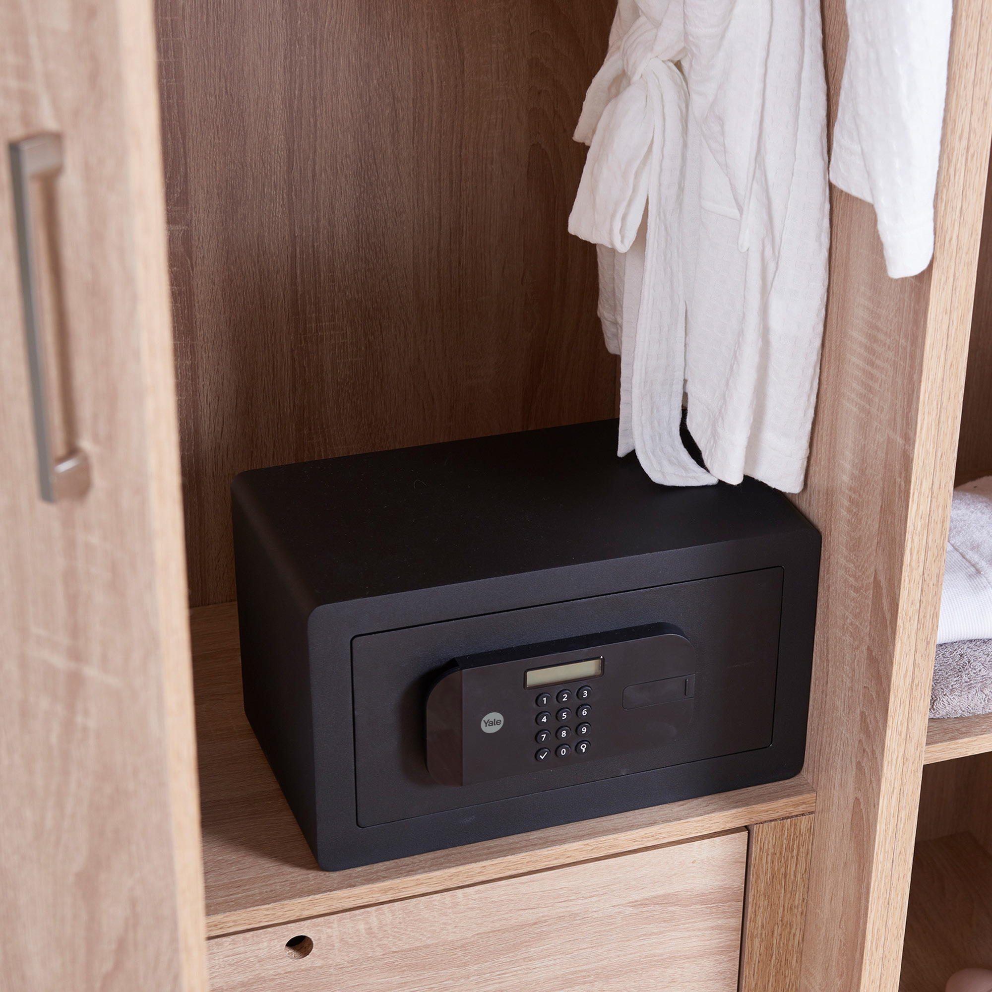 Ein schwarzer Safe mit Tastatur in einem Holzschrank. Ein weißer Bademantel hängt darüber, ein Handtuch rechts.