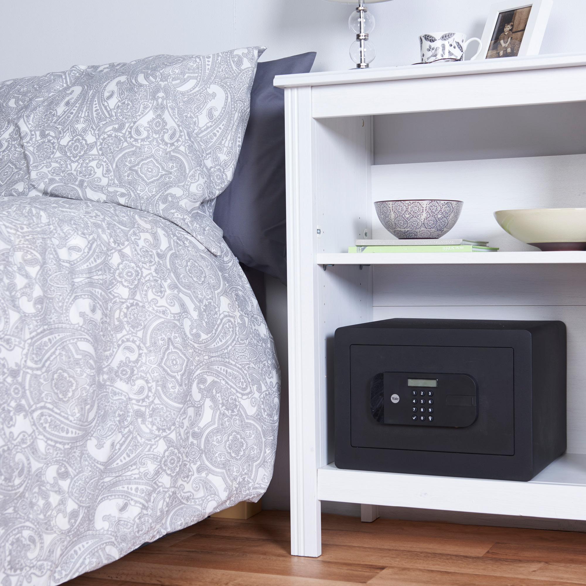 Ein Bett mit Paisley-Bettwäsche neben einem weißen Nachttisch. Auf dem unteren Regal befindet sich ein schwarzer Safe.