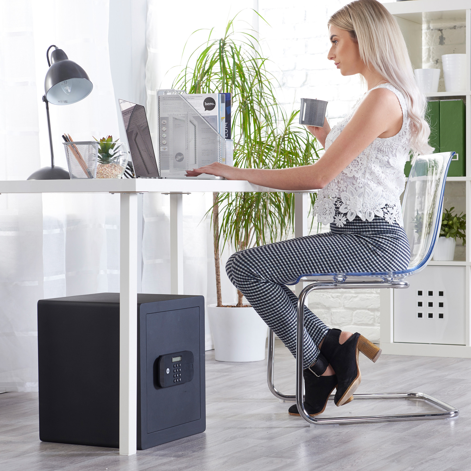 Blonde Frau sitzt an einem Schreibtisch mit Laptop und hält eine Tasse. Ein Safe steht auf dem Boden, eine Pflanze ist zu sehen.