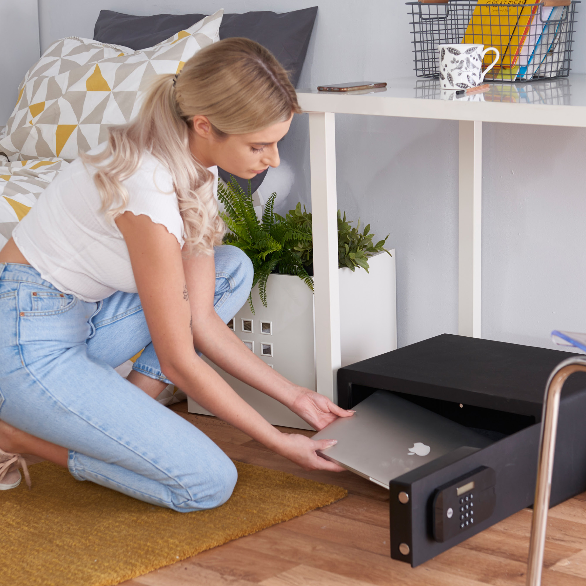 Eine Frau legt einen Laptop in einen schwarzen Safe unter einem weißen Schreibtisch. Der Raum hat Pflanzen, ein Bett und eine Kaffeetasse.