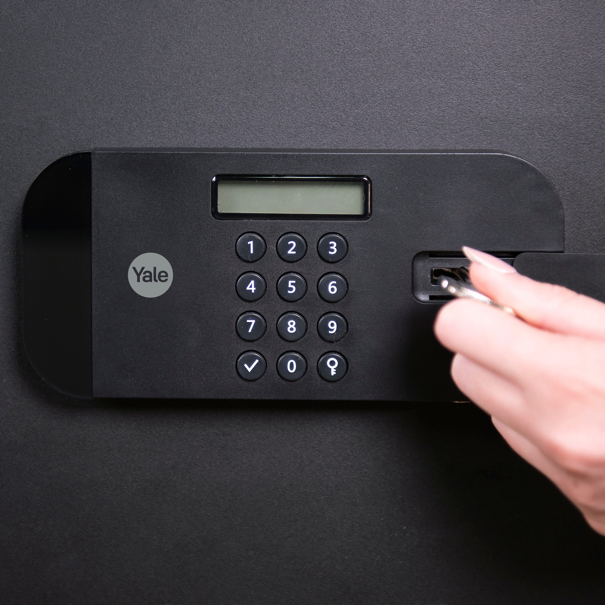 Eine Hand steckt einen Schlüssel in einen schwarzen Safe. Der Safe hat eine Tastatur und ein Display. Die Marke Yale ist sichtbar.