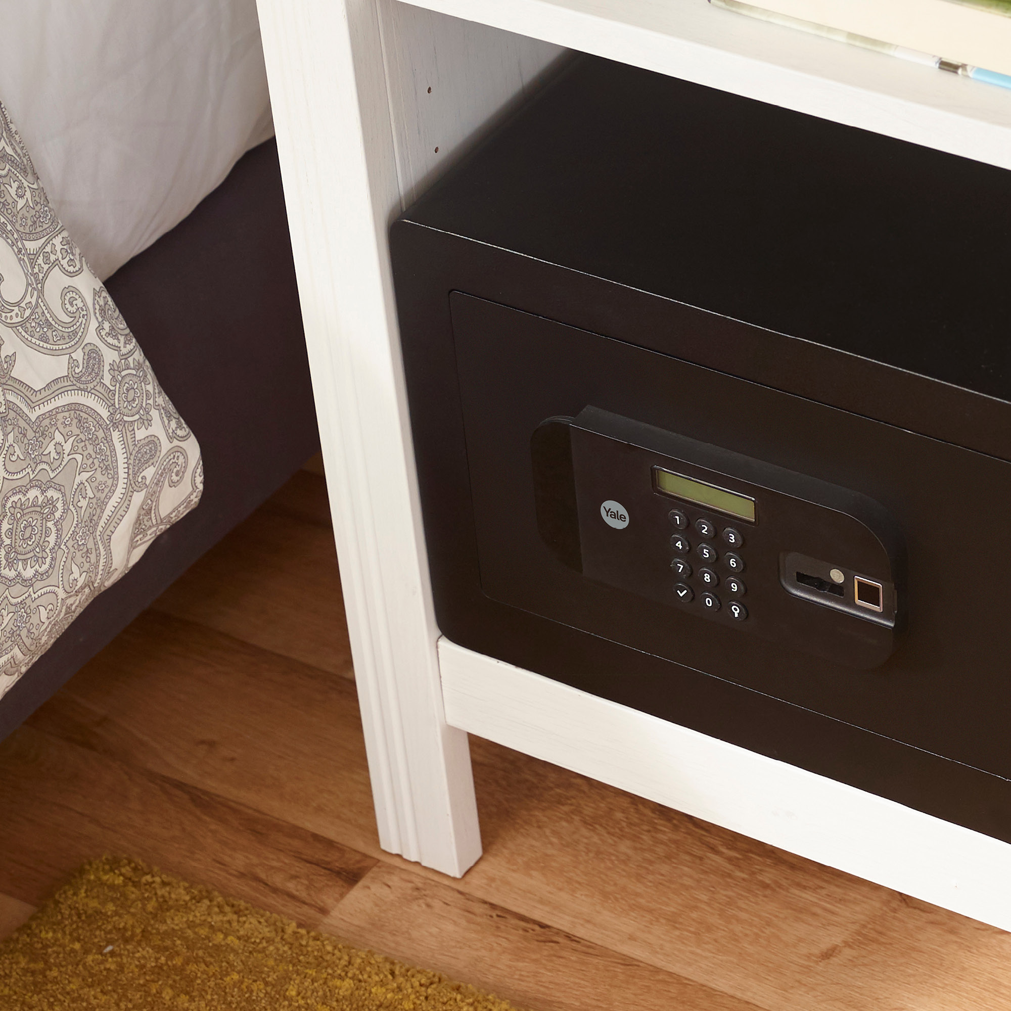 Ein schwarzer Safe mit Tastatur und Yale-Logo steht in einem weißen Nachttisch. Links ein Bett mit gemusterter Bettwäsche.